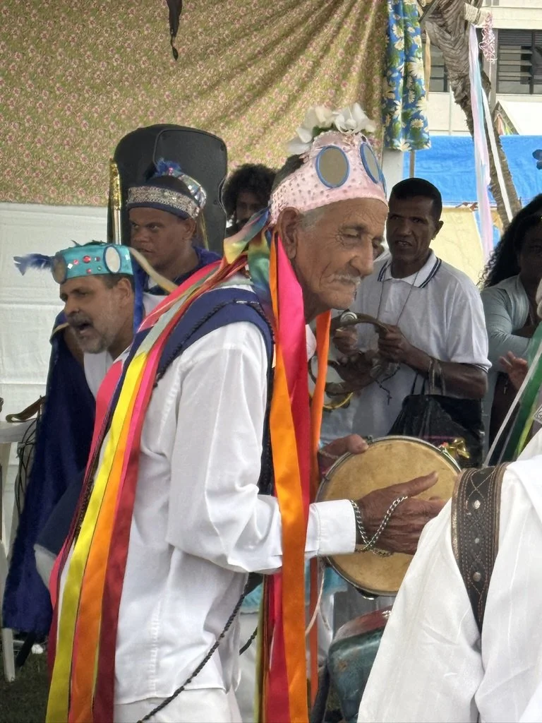 An elderly man wearing a pink hat with colorful ribbons and round sunglasses on top, playing a tambourine during a cultural celebration or festival. Several other people in festive attire and stage decorations are visible in the background.