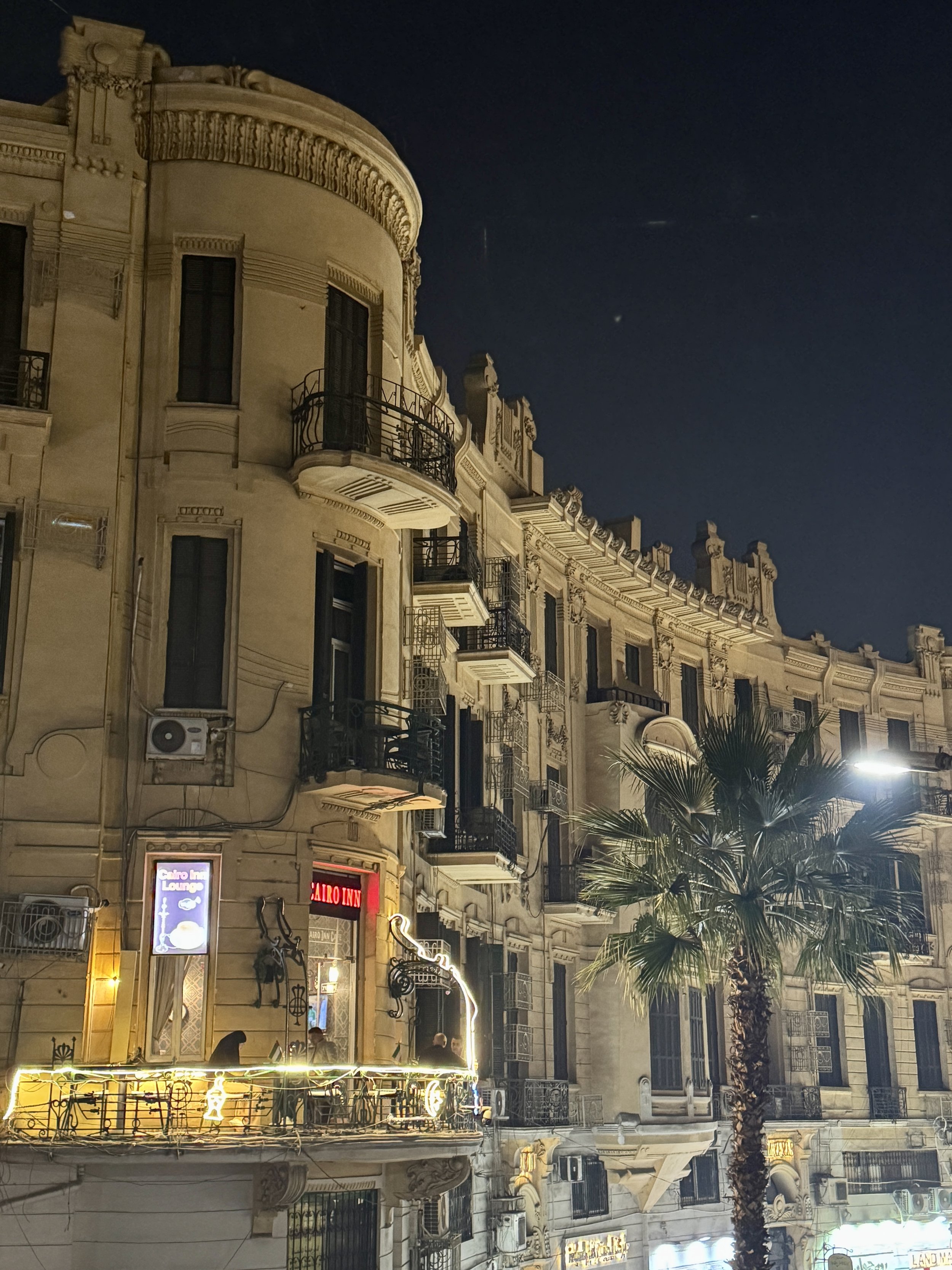 Night view of an ornate, European-style building with balconies, decorative molding, and illuminated signs at street level. A palm tree is in the foreground.