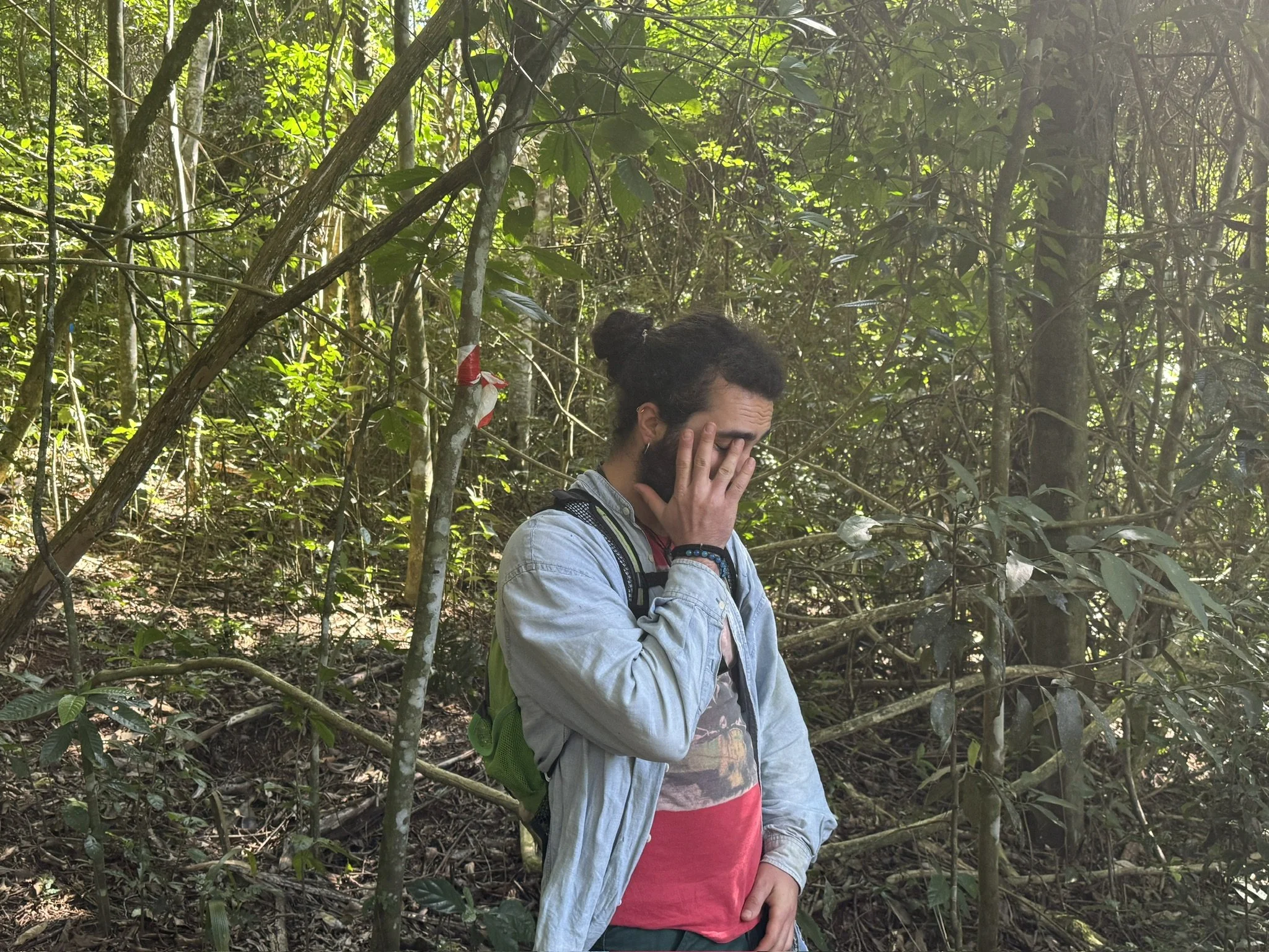 A person with curly dark hair and a beard, wearing a denim jacket and red t-shirt, covers their face with both hands in a dense forest with tall trees and green foliage.