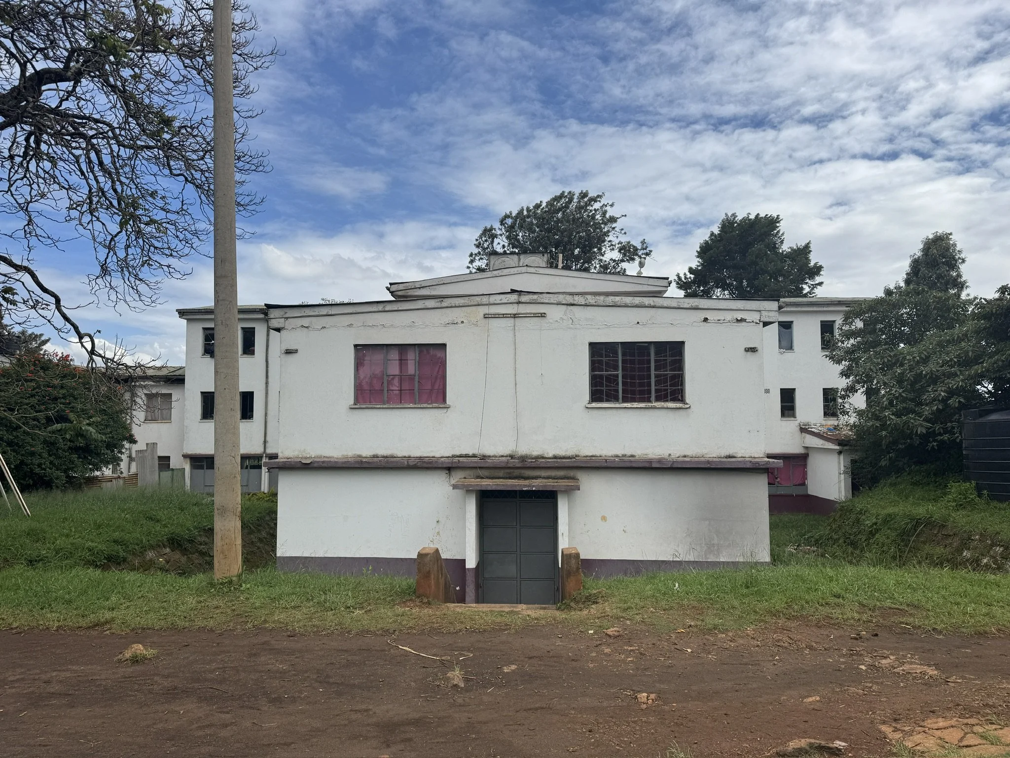 A white two-story building with multiple windows, some covered with pink curtains, and a gray door at the bottom center. The building is set against a partly cloudy blue sky, with trees on either side and around it. The foreground shows a patch of di
