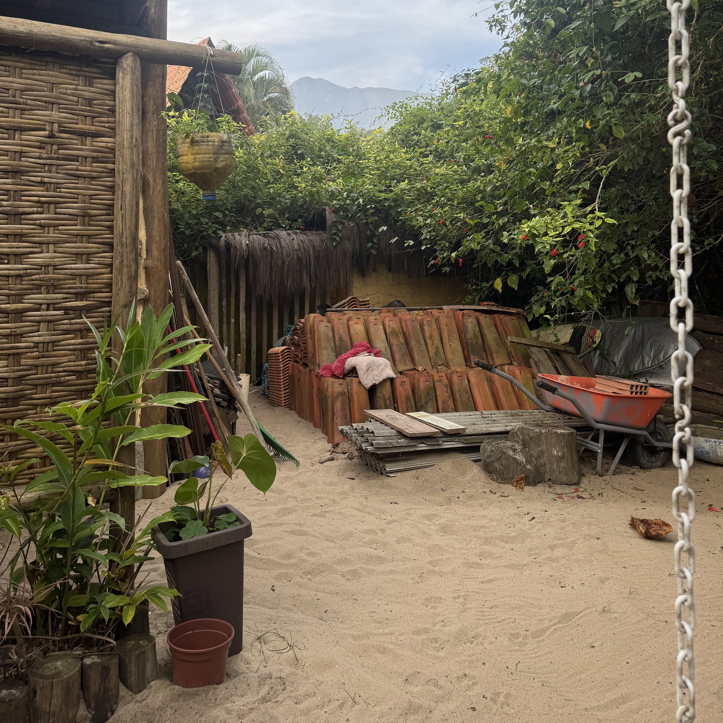View of a rustic outdoor area with sand ground, potted plants in the foreground, stacked clay tiles, a wheelbarrow, and lush green trees and shrubs in the background. Mountain peaks are visible in the distance under a cloudy sky.