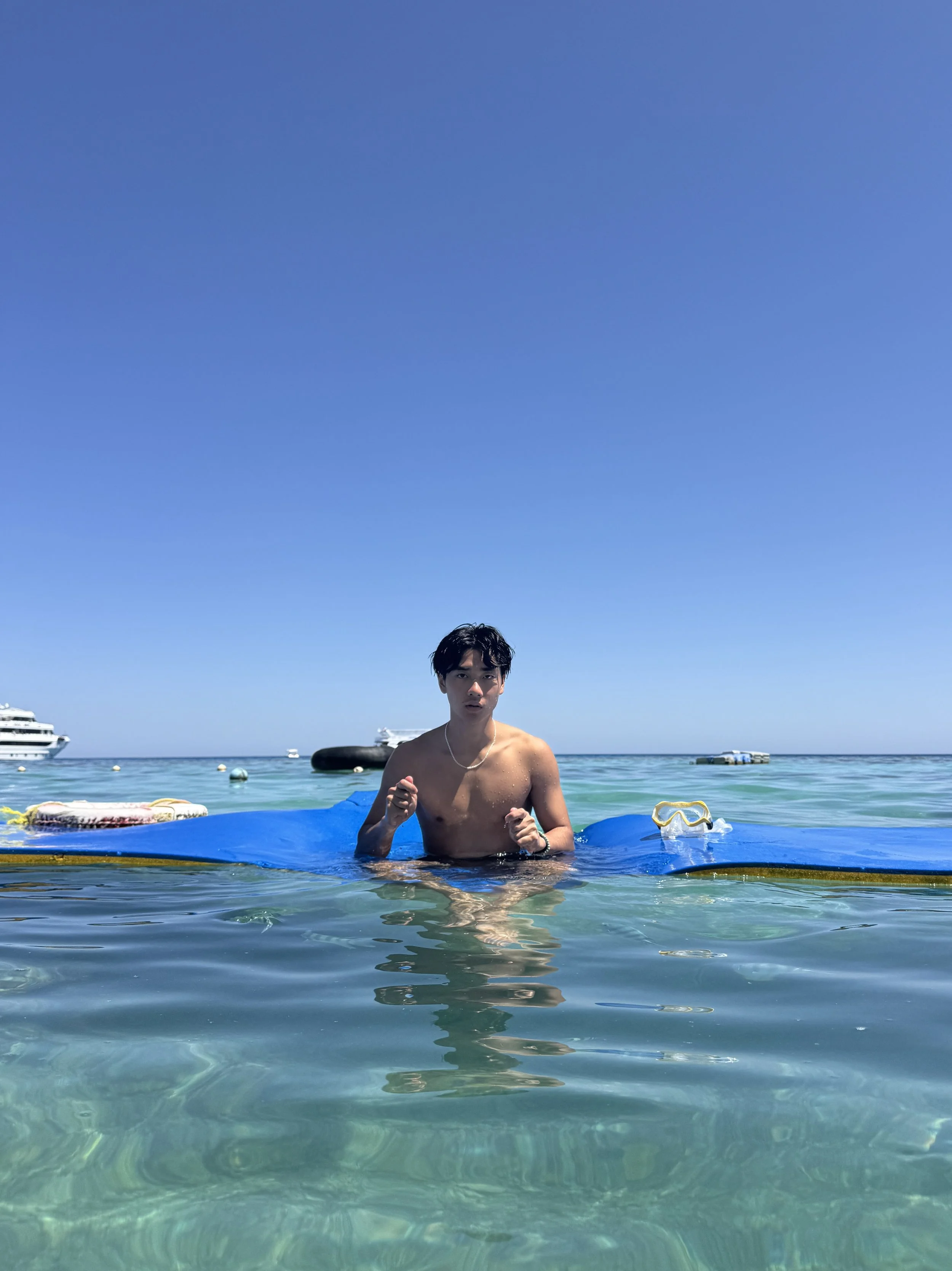 A young man with dark hair and a necklace stands in shallow clear water at the beach, with boats and yachts in the background and a bright blue sky overhead.