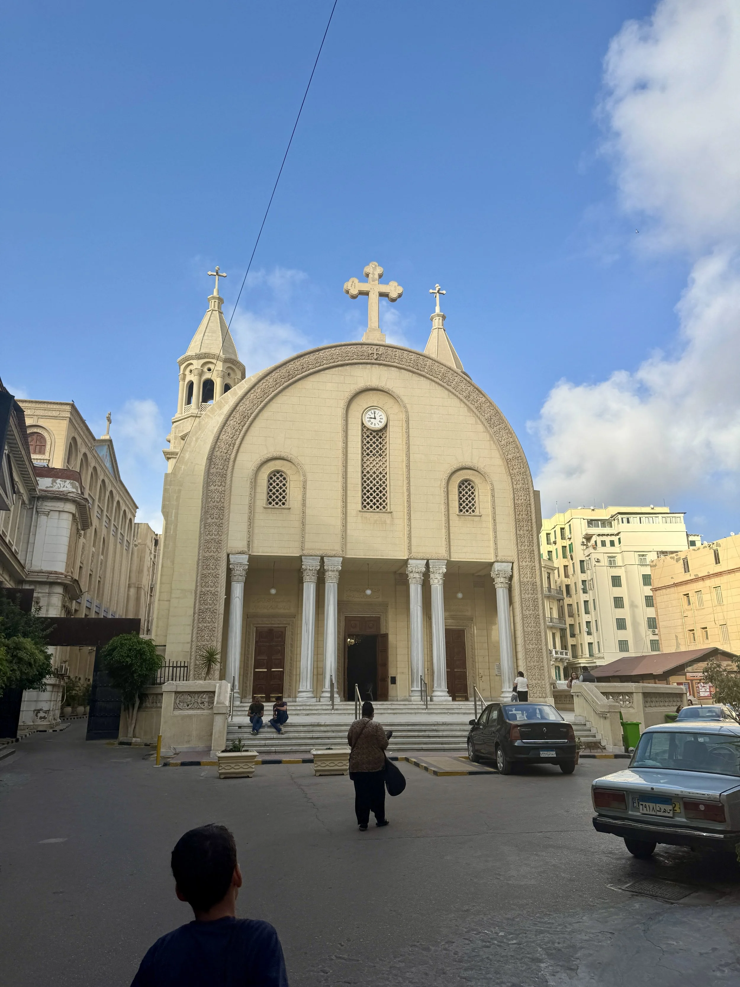 A church with a large cross on top, featuring two smaller towers with crosses, and a clock on the front facade. Several people are sitting and standing outside, with cars parked in the surrounding area under a partly cloudy sky.