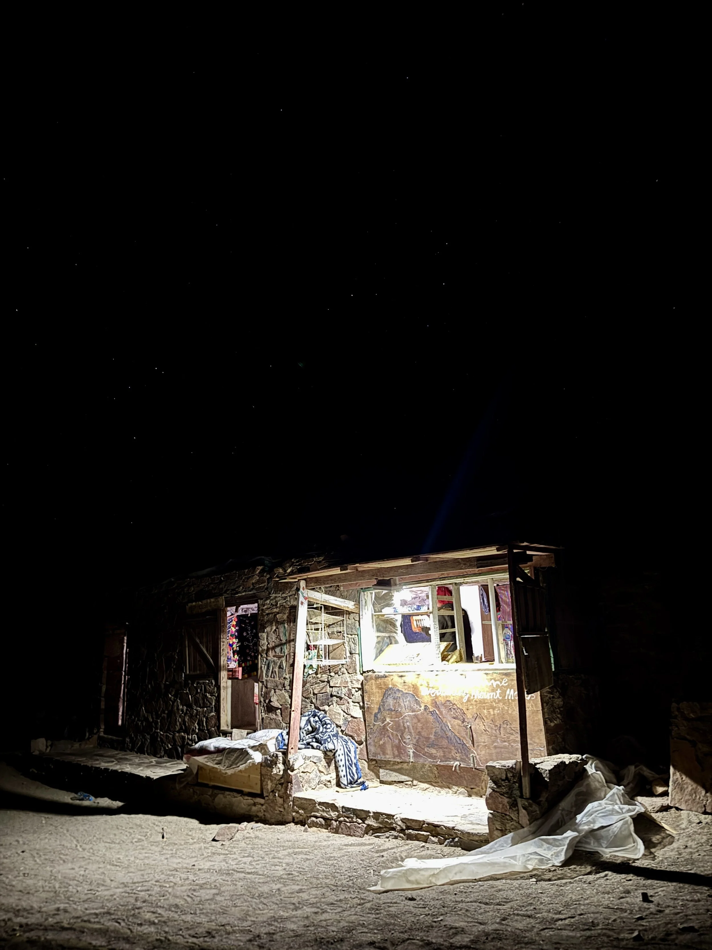 Nighttime view of a partially constructed or damaged stone building or shelter with a large open window or doorway illuminated from the inside. The scene shows various construction materials, debris, and fabric on the ground and inside, with a sign i