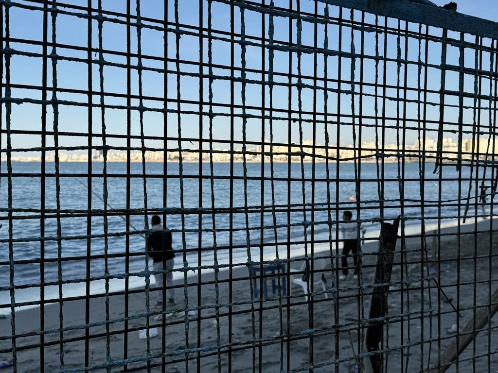 View of a beach seen through a wire fence, with two people walking along the shoreline and a boat on the water, under a clear sky.