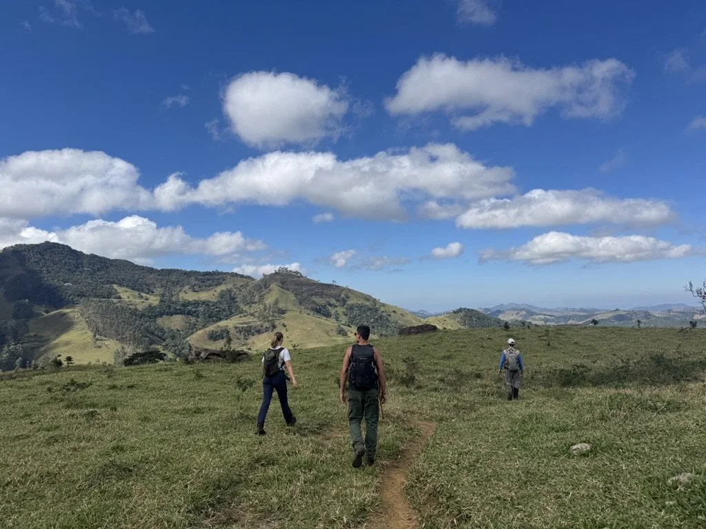 Three hikers walking on a trail in a grassy open field under a blue sky with clouds, with hills and mountains in the distance.
