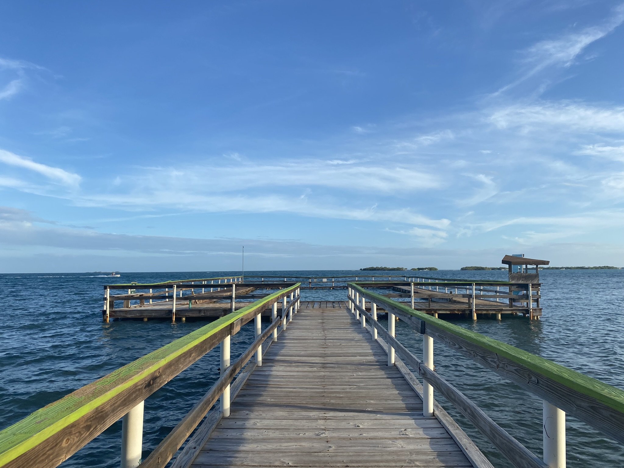 A wooden pier extending into a body of water with railing, under a blue sky with some clouds, and a small island visible in the distance.