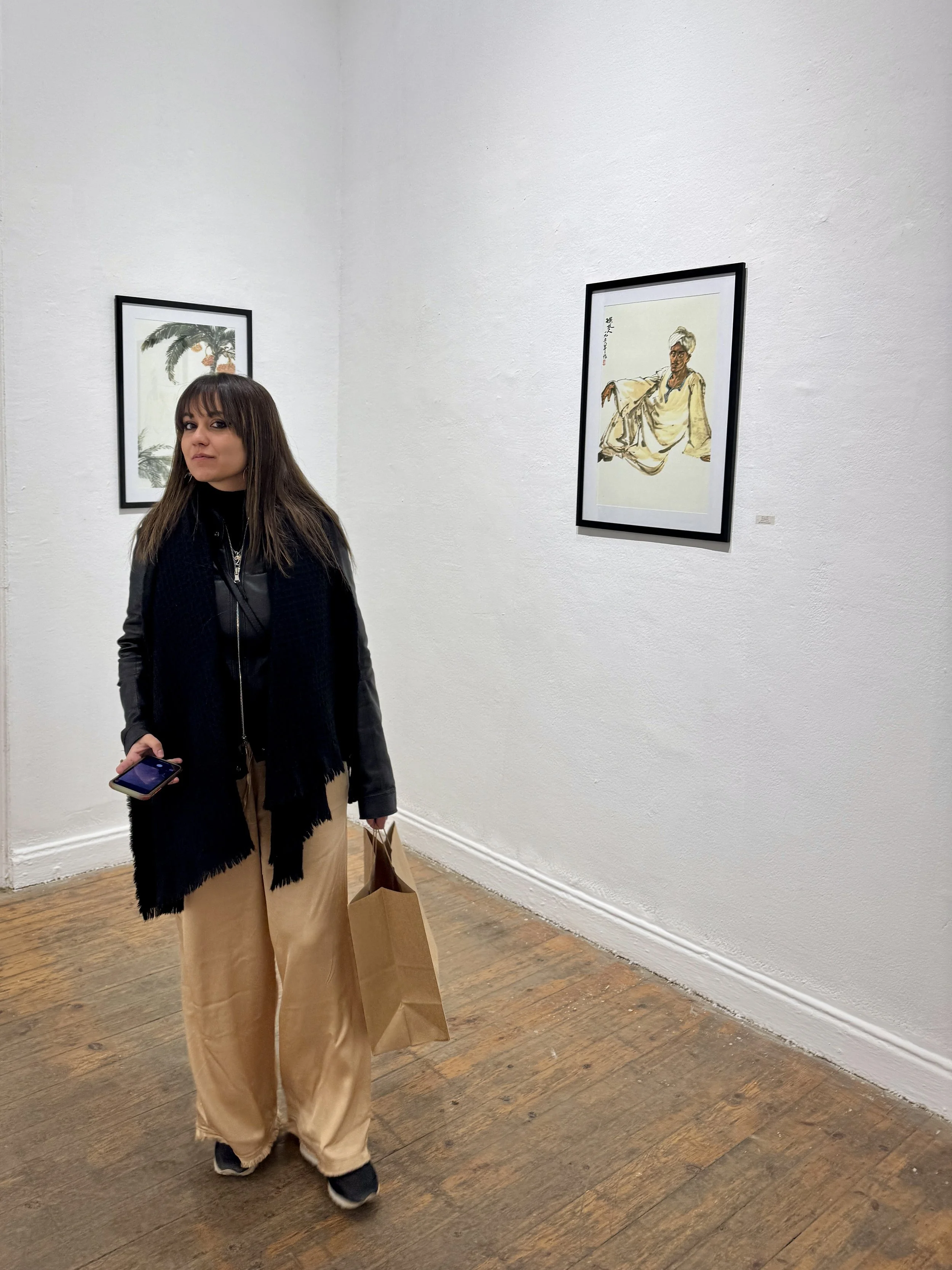 A woman with brown hair, wearing a black jacket and beige pants, stands in an art gallery. She is holding a phone in her right hand and shopping bags in her left hand. Two framed artworks are visible on the white wall behind her, one depicting a pers