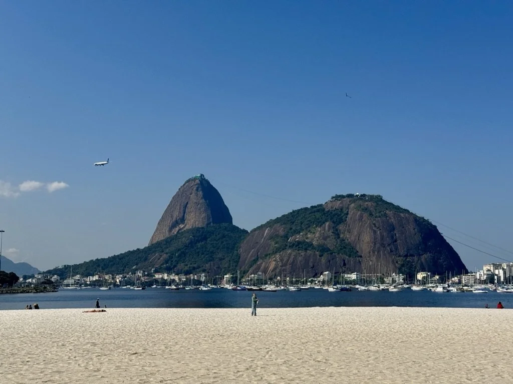 View of Sugarloaf Mountain and Corcovado Mountain in Rio de Janeiro, Brazil, seen across a sandy beach with some people walking and relaxing, and boats anchored in the water.