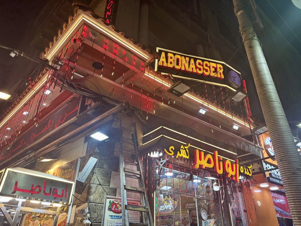 Night view of a restaurant storefront with bright neon signs in Arabic and English, including the words 'ABONASSER.' The signage is decorated with red and yellow lights.
