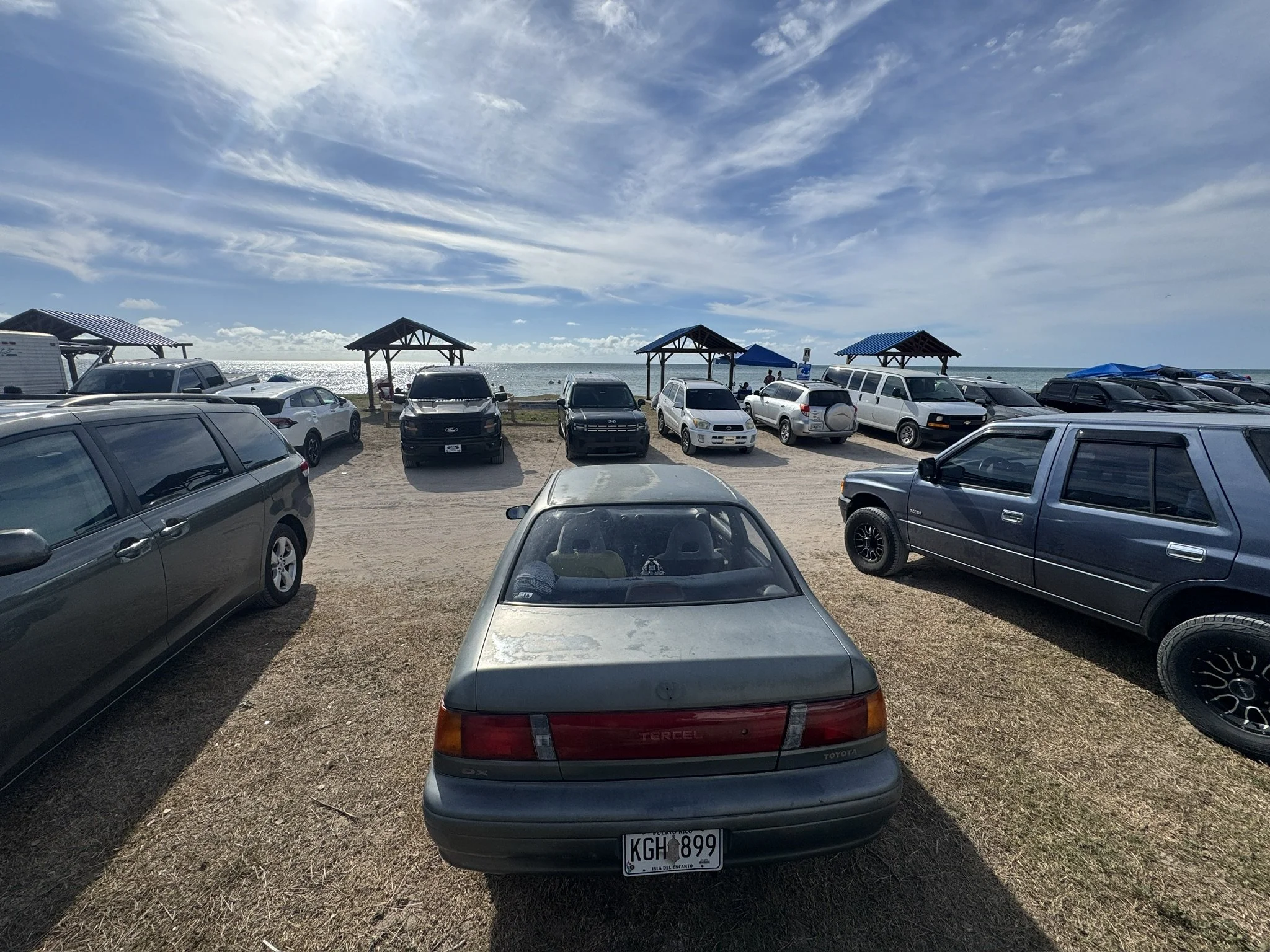 A parking lot near the beach with several cars parked on a dirt surface and small shaded structures, with the ocean and a partly cloudy sky in the background.