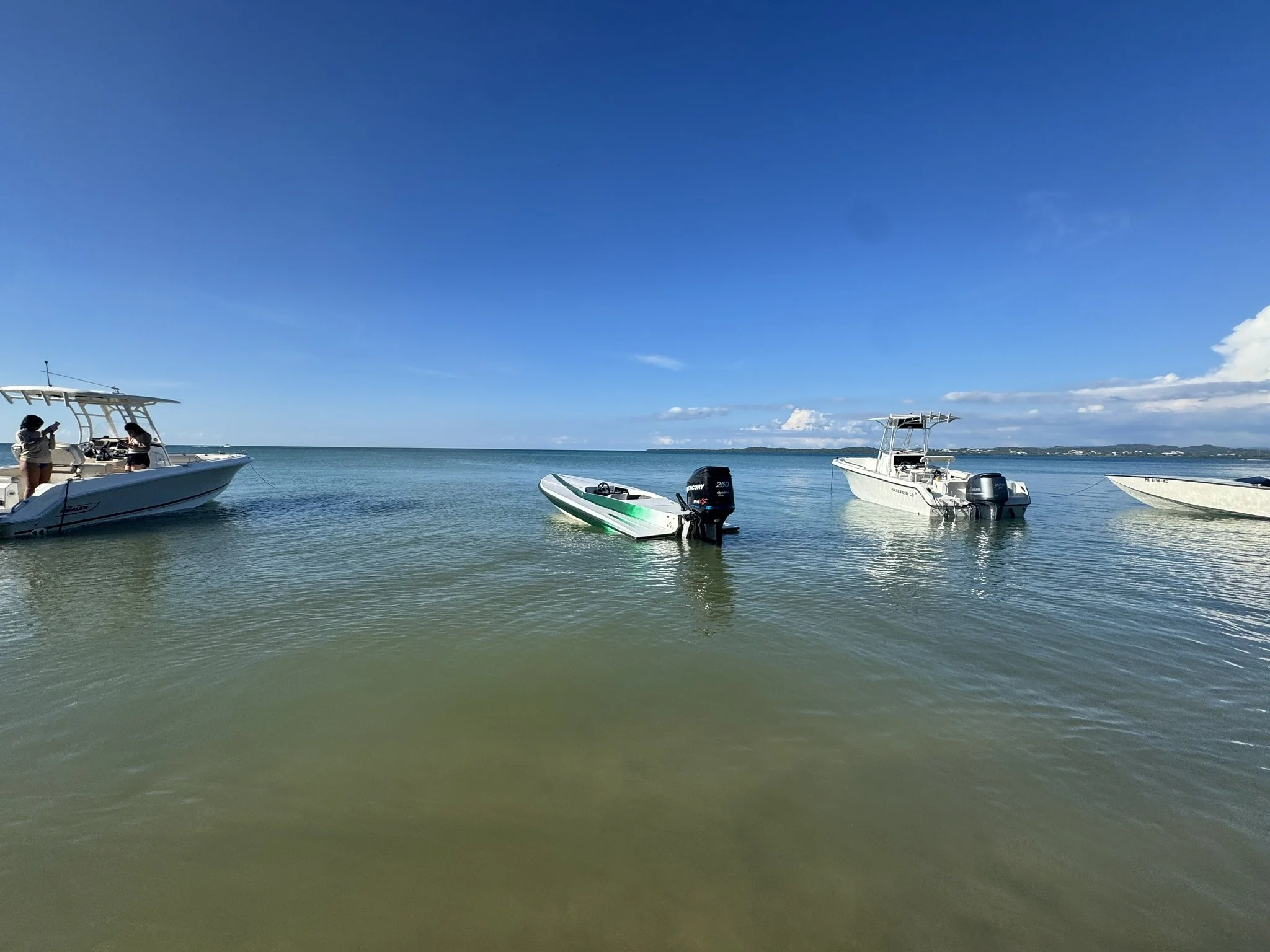 Several boats float on calm water near a pier with two people standing on one boat. The background shows a distant shoreline with a clear blue sky and some clouds.