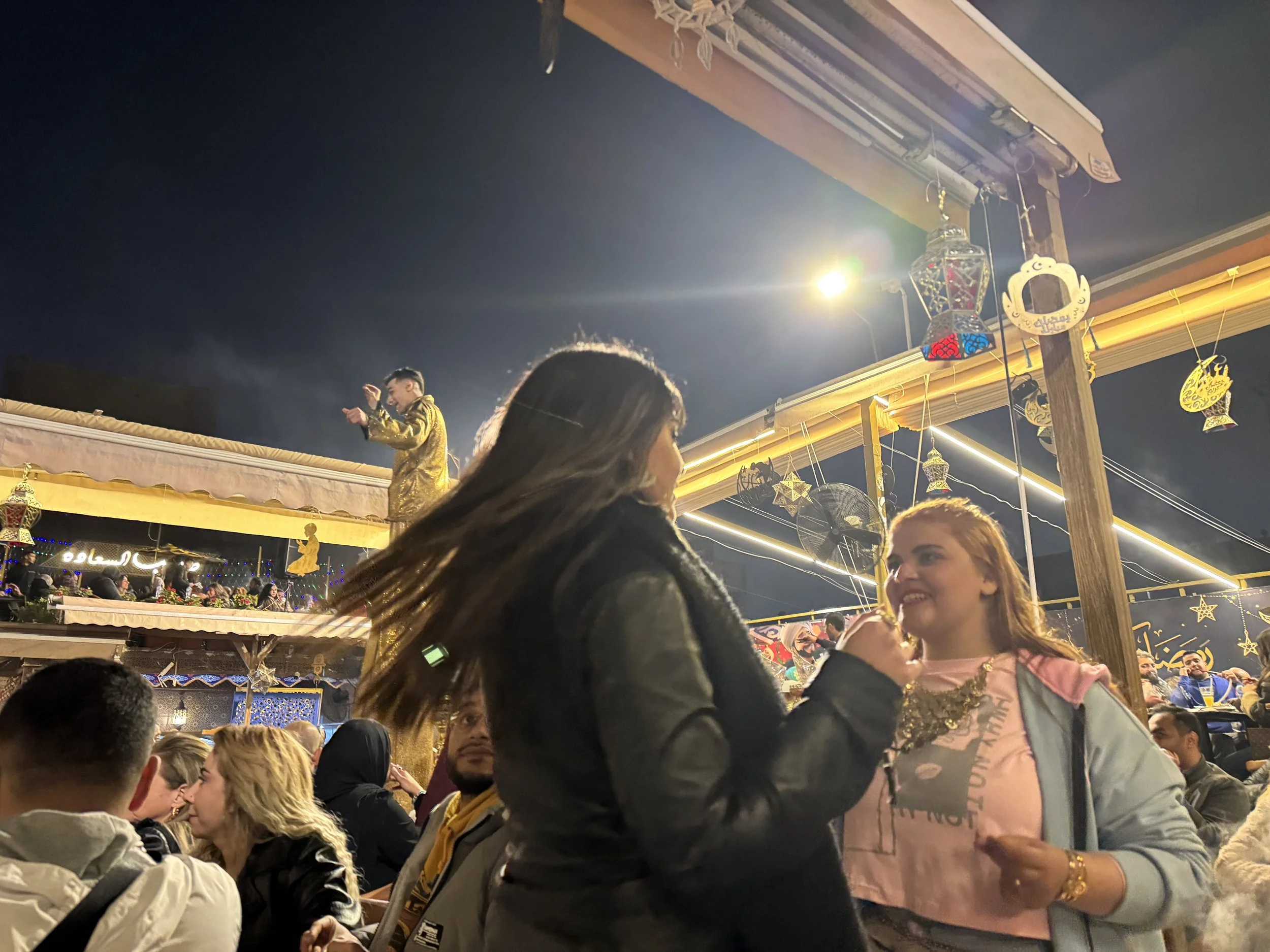 A crowded outdoor celebration at night with festive decorations, including hanging lanterns and ornaments. People are socializing and enjoying the event, and a performer in a gold outfit is present on a stage or platform.