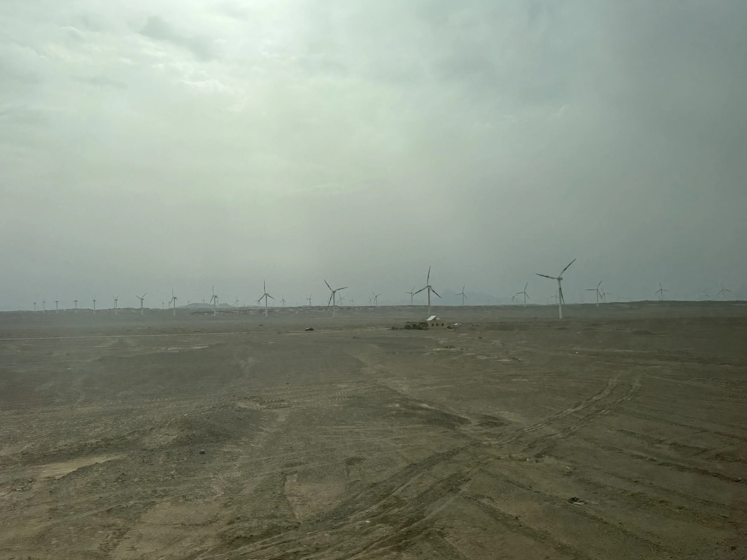 A wind farm with multiple wind turbines visible under a cloudy sky, situated in a flat, open landscape with dirt ground.