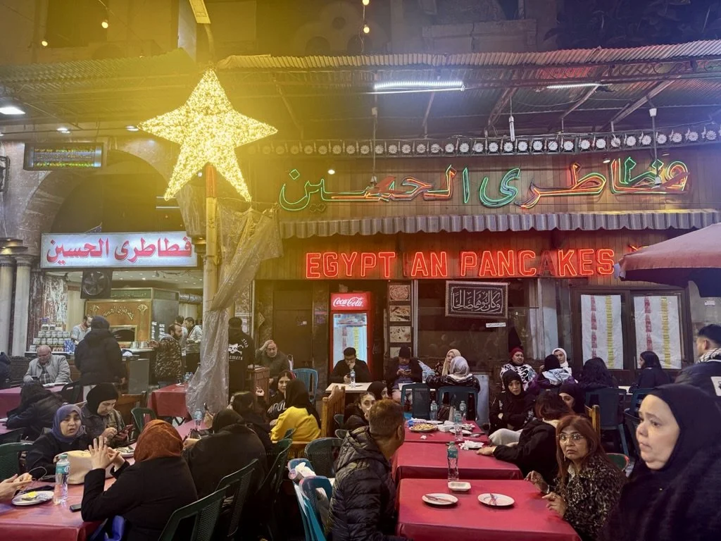 Indoor restaurant with people seated at tables, neon sign reading 'Egyptian Pancakes' and Arabic signage, large star decoration, and festive lighting.