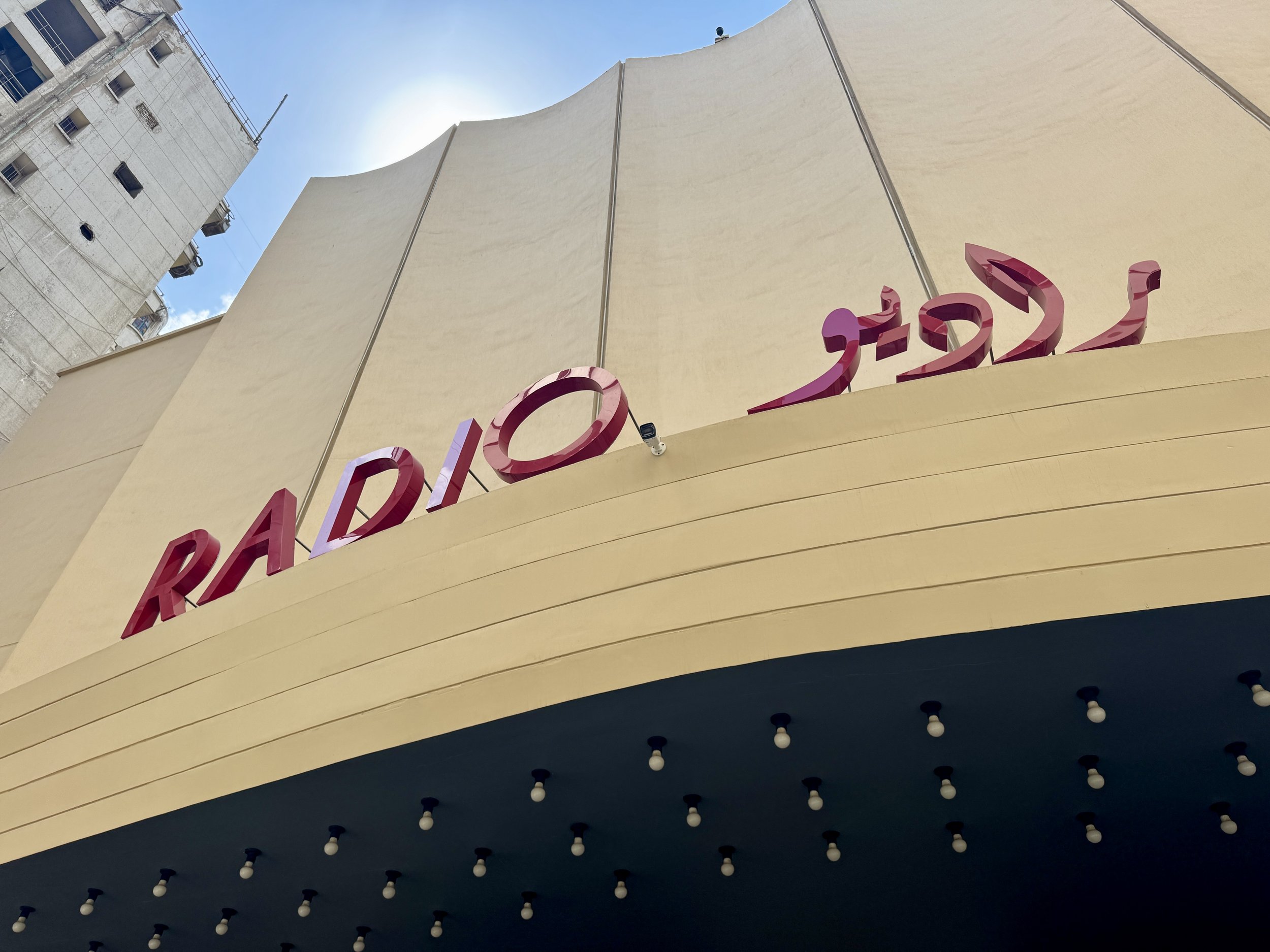 Low-angle view of a building with a beige curved structure and red signage, against a bright blue sky with the sun shining.