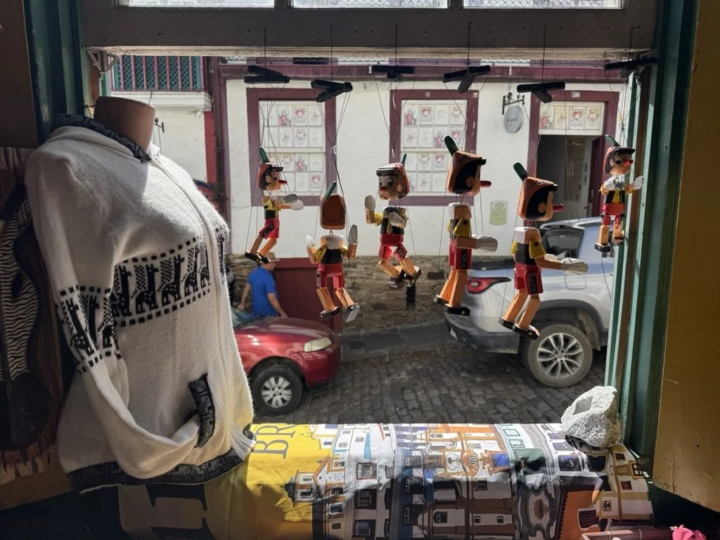 View of a shop window display featuring wooden Pinocchio puppets hanging in front of the window, with a mannequin head draped in a white sweater on the left, and a colorful tablecloth with a cityscape pattern on the window sill.
