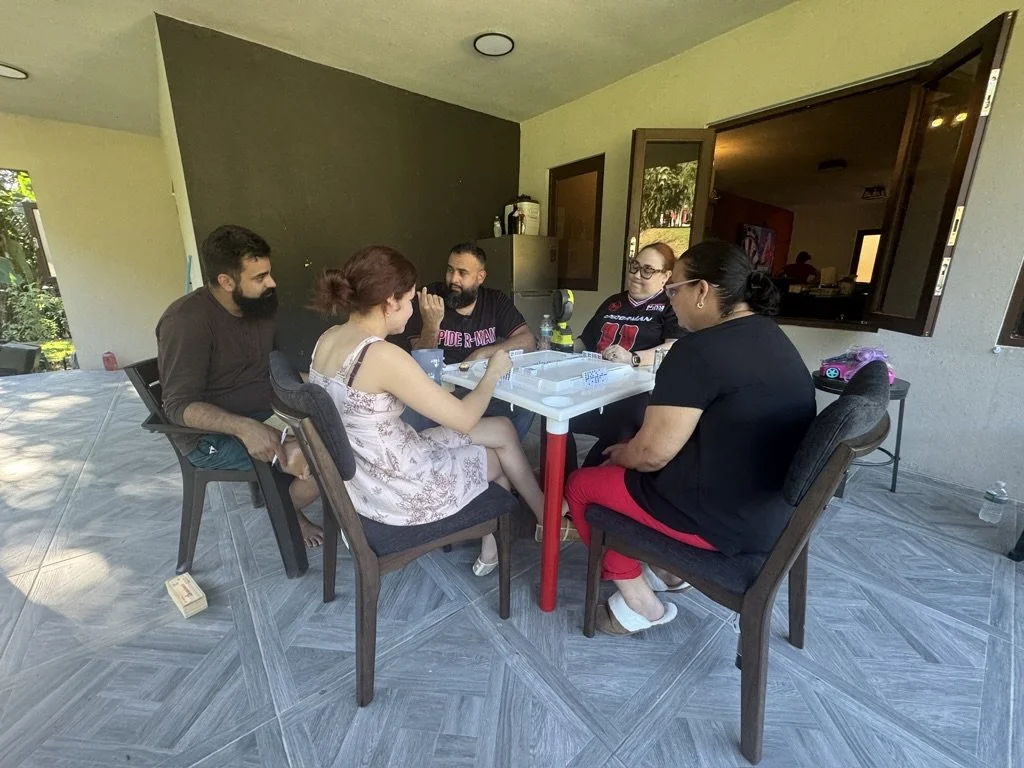 Six people sitting around a white table on a covered patio, engaged in conversation, with open windows showing a kitchen interior.