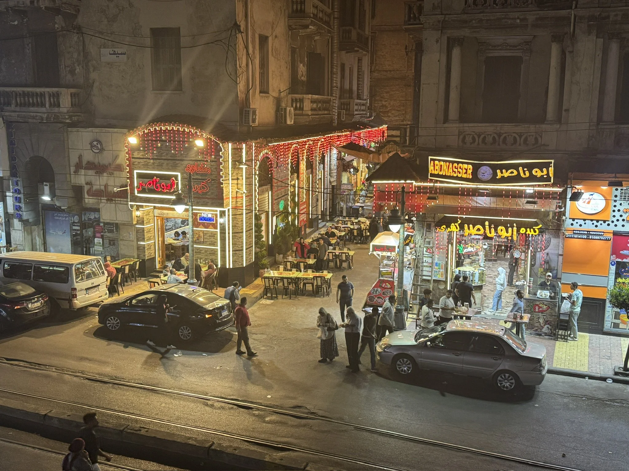 Night-time street scene with people walking and sitting outside cafes, parked cars along the street, and illuminated signs in Arabic and English, with old buildings in the background.