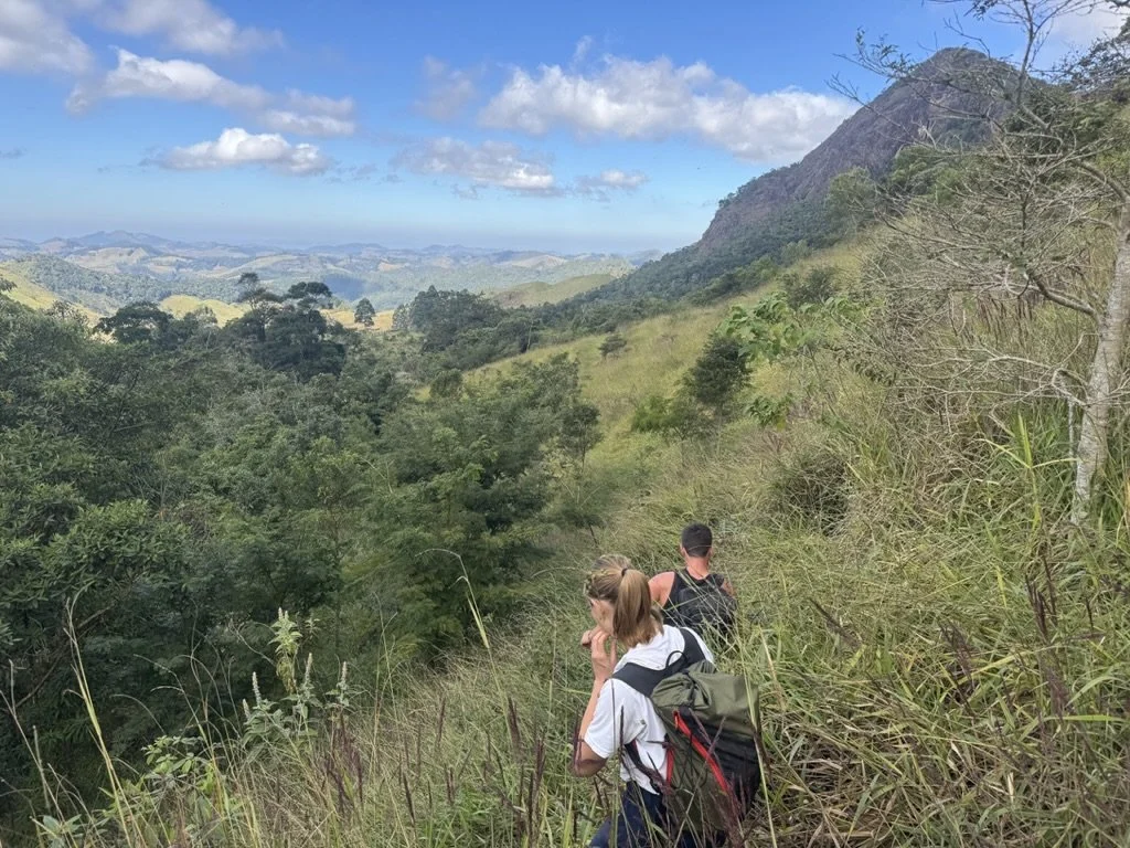 Three hikers trekking through a lush, green hillside trail with a mountainous landscape and partly cloudy sky in the background.