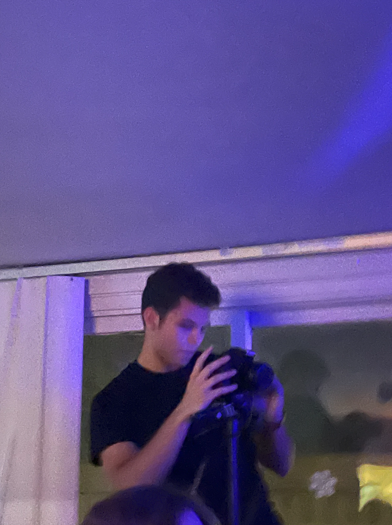 A young man with dark hair is taking a selfie in a mirror using his phone. The room is lit with dim, purple-blue lighting. Part of another person's head is visible at the bottom of the photo.