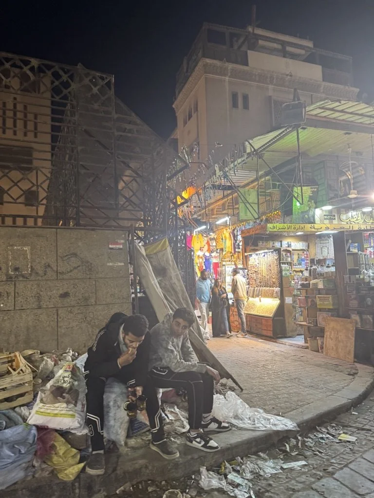 Two young men sit on a curb at night in front of a street market, with garbage around them, and a busy shop with colorful items and bright lights behind them.