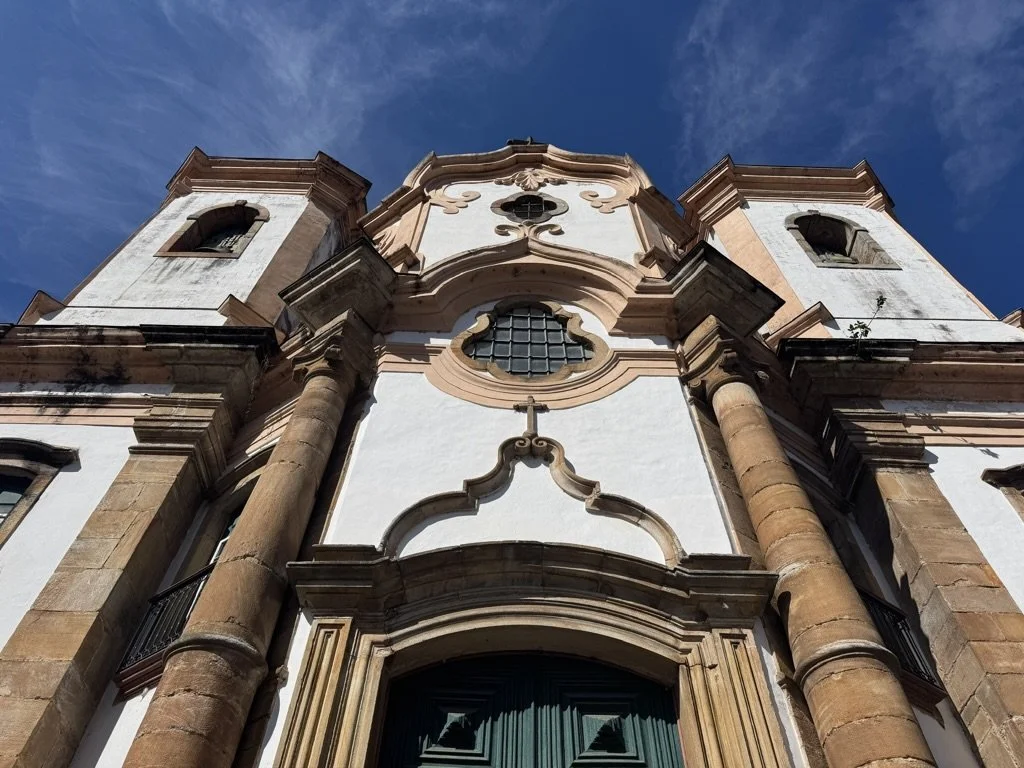 Low-angle view of a historic church with baroque architecture, featuring tall bell towers, ornate window frames, and decorative stucco details against a blue sky.