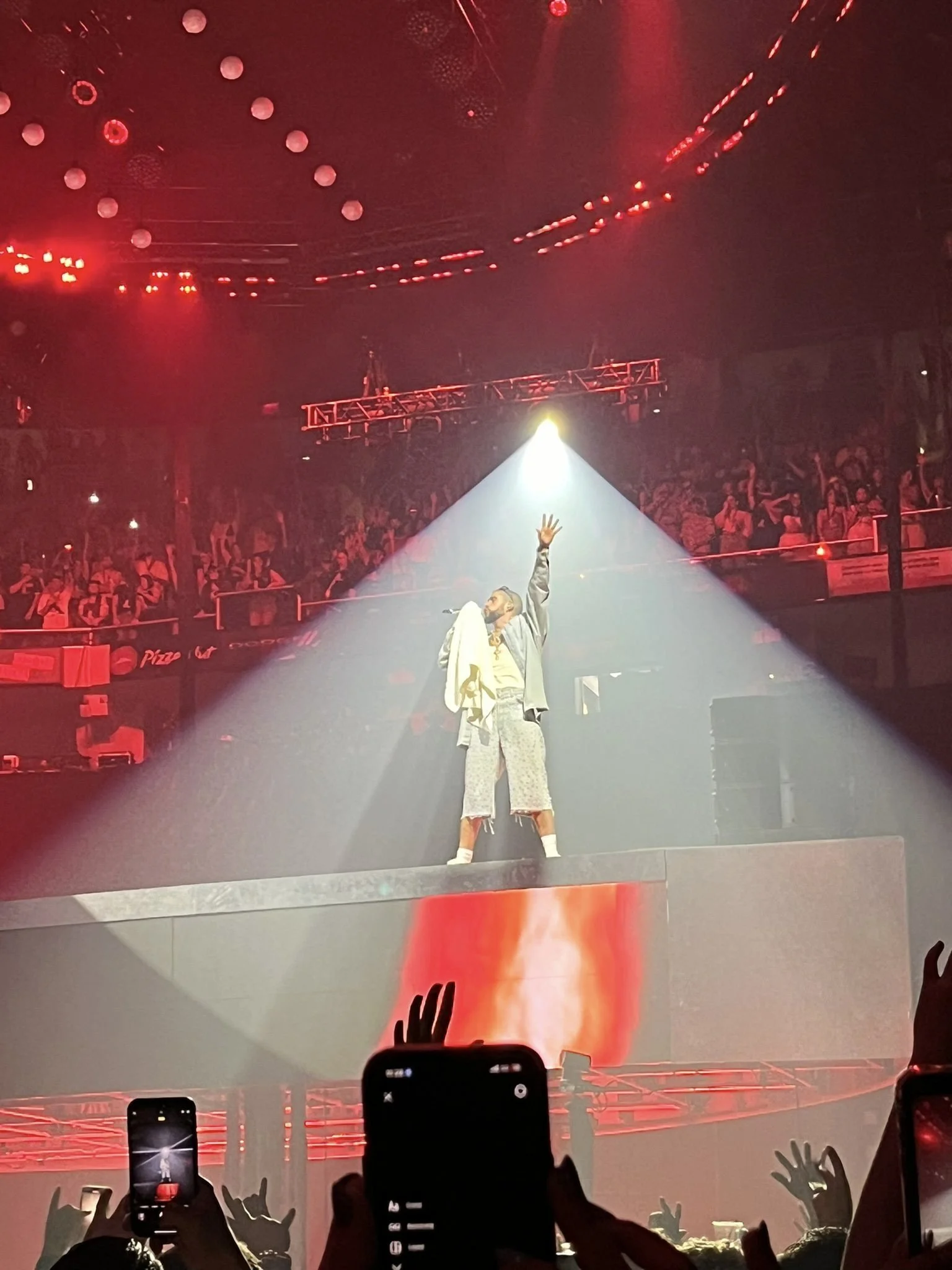 A performer on stage under a spotlight during a concert, with an audience raising hands and taking photos, red stage lights, and hanging balloons and decorations overhead.