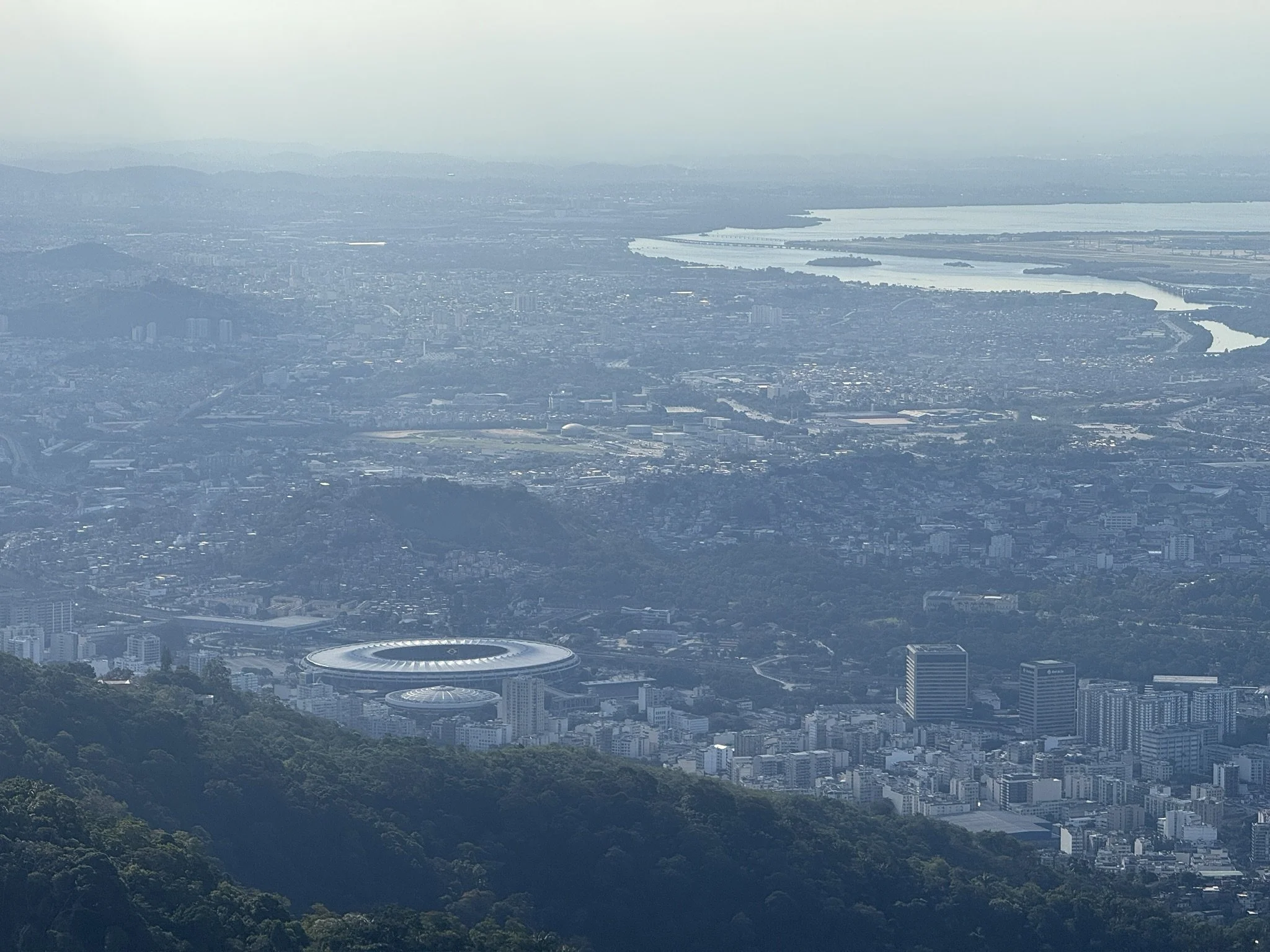 Aerial view of a cityscape with a stadium, water bodies, and mountains in the distance.