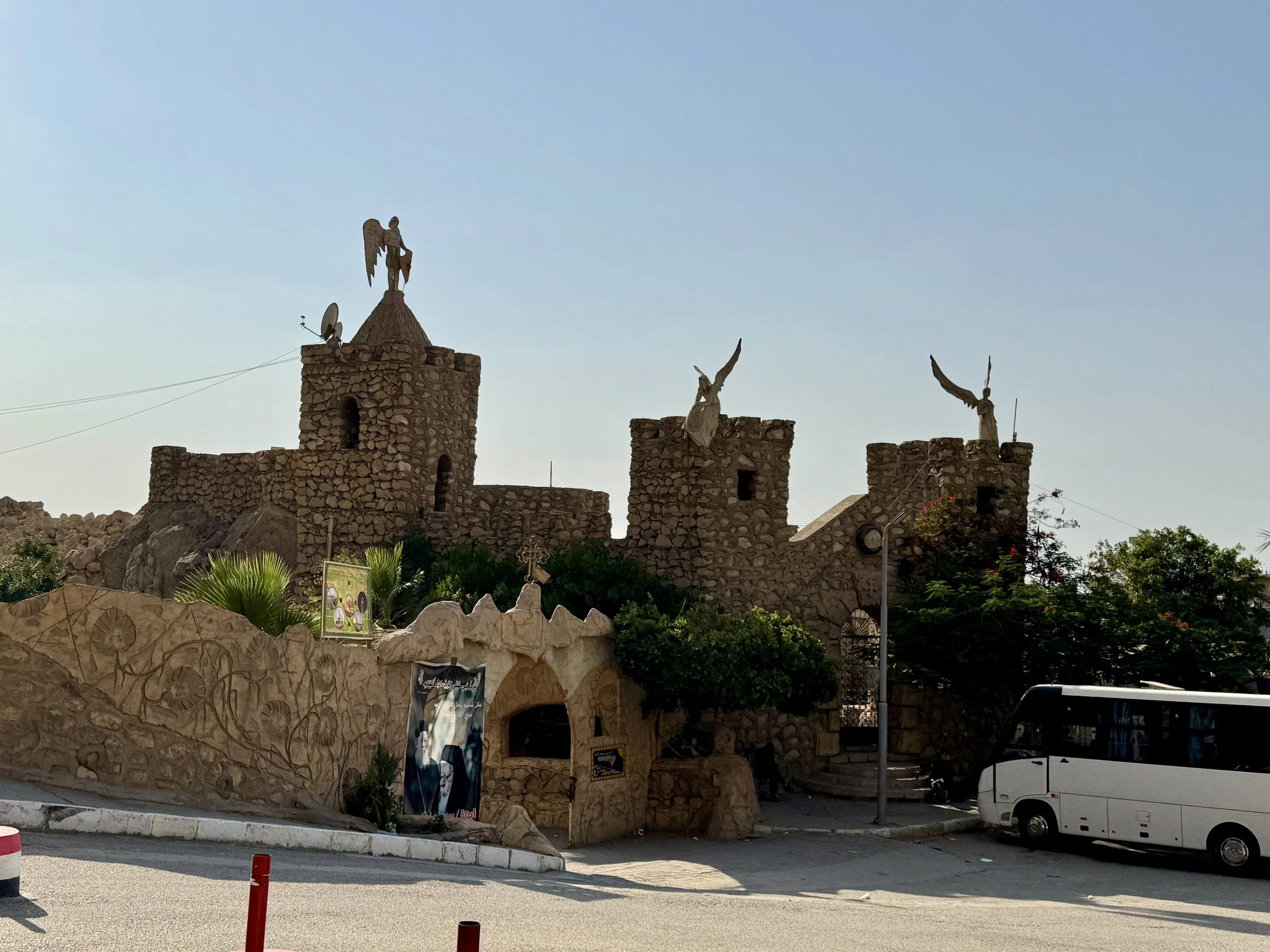 Stone castle-like structure with several statues on top, including angel figures with wings, against a clear blue sky. There is a satellite dish and wires attached to the structure, with greenery and a bus parked nearby.