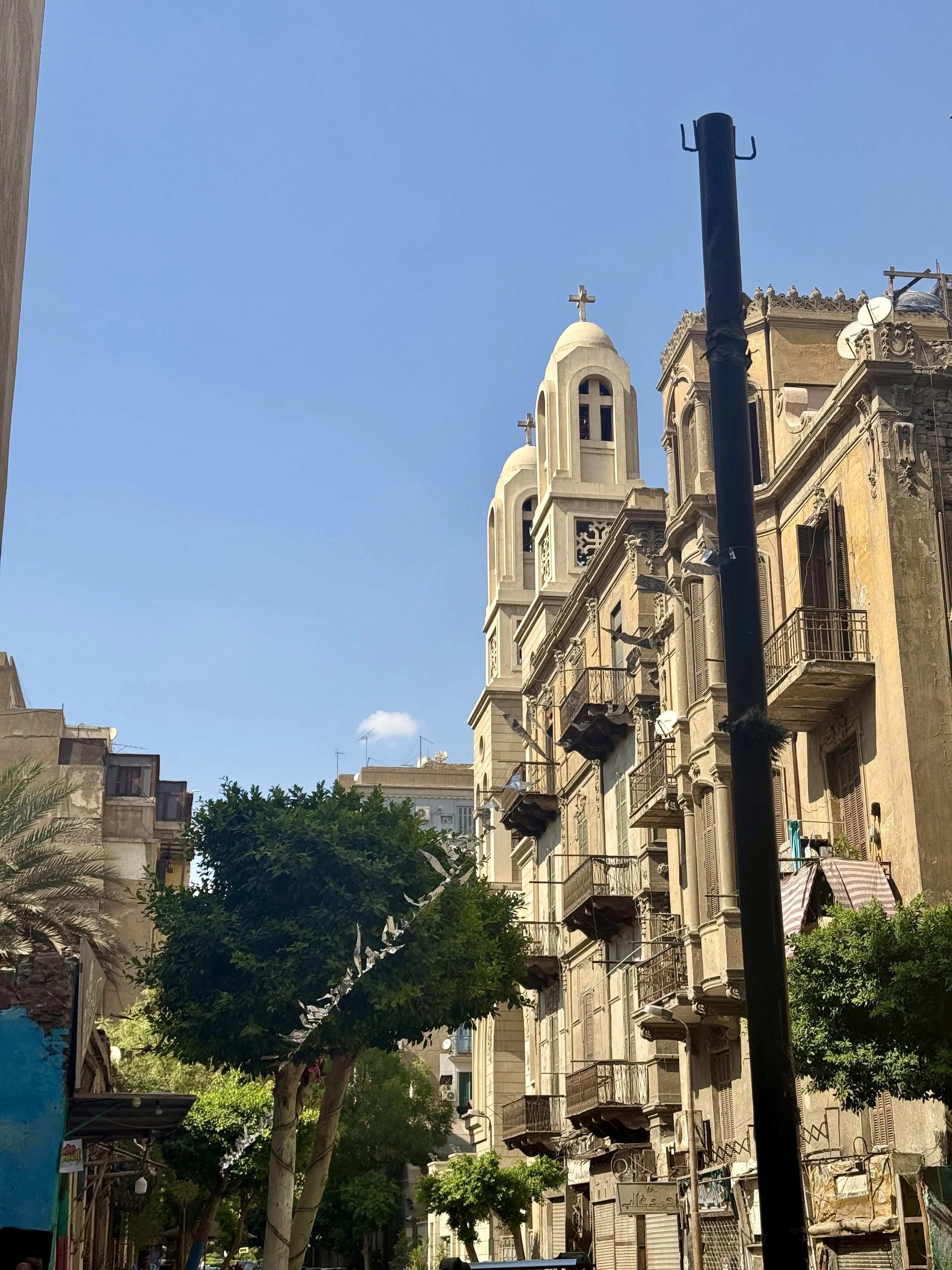 City street with old buildings, balconies, trees, and a church with a cross on top under a blue sky.
