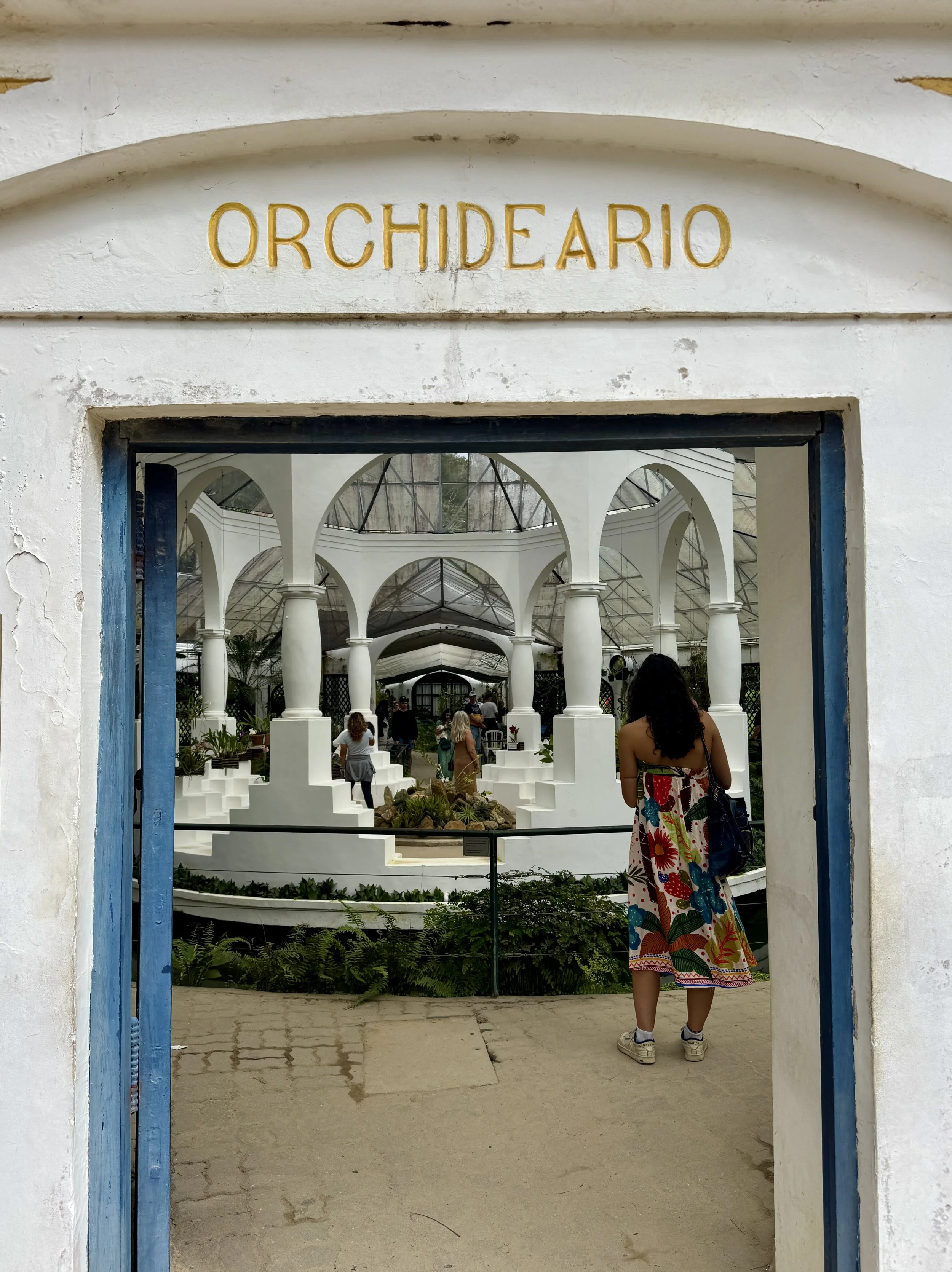 Entrance to a greenhouse or conservatory named 'Orquideario' with white arched architecture and people inside, including a woman in a colorful dress standing at the entrance.