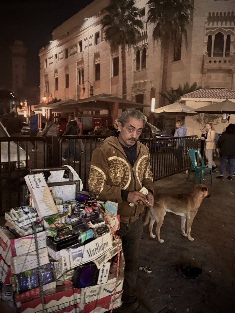 A man standing next to a cart of cigarette packs and lighters on a city sidewalk at night. A dog stands nearby, with people and historic buildings in the background.