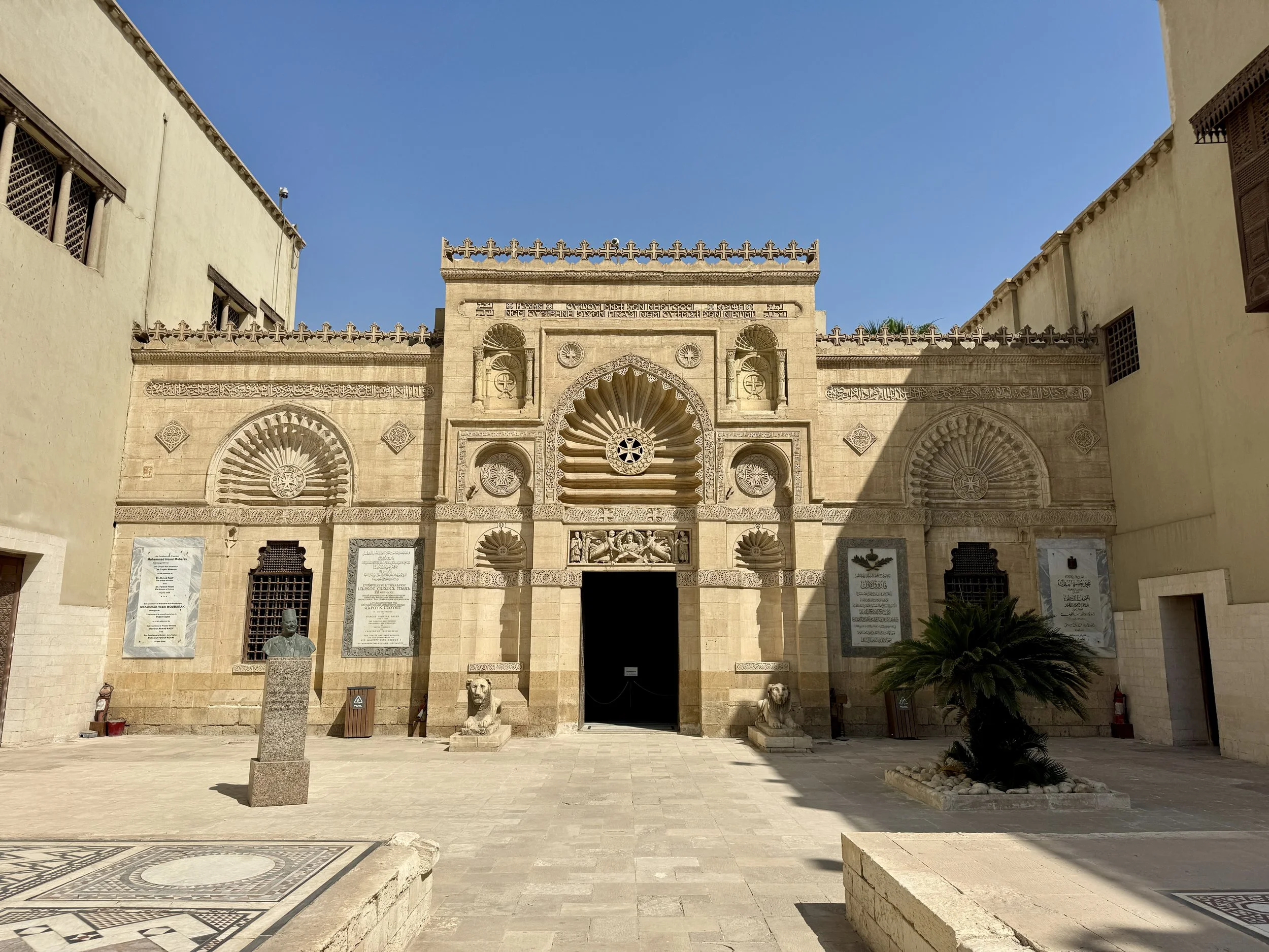 Front view of a historical building with intricate stone carvings, statues, and inscriptions, flanked by two lion sculptures at the entrance, under a clear blue sky.