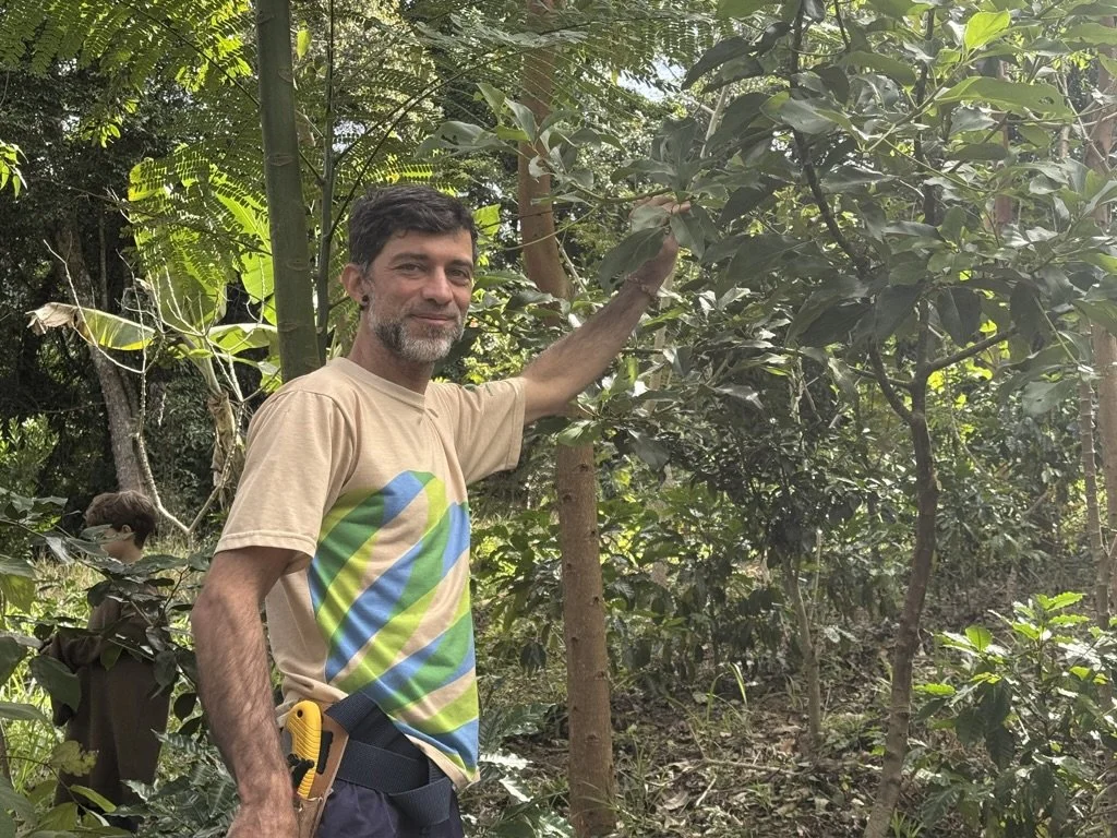 A man with short dark hair and a beard standing in a lush forest, touching a tree with his right hand, wearing a beige t-shirt with a colorful circle design, and a tool attached to his waistband.