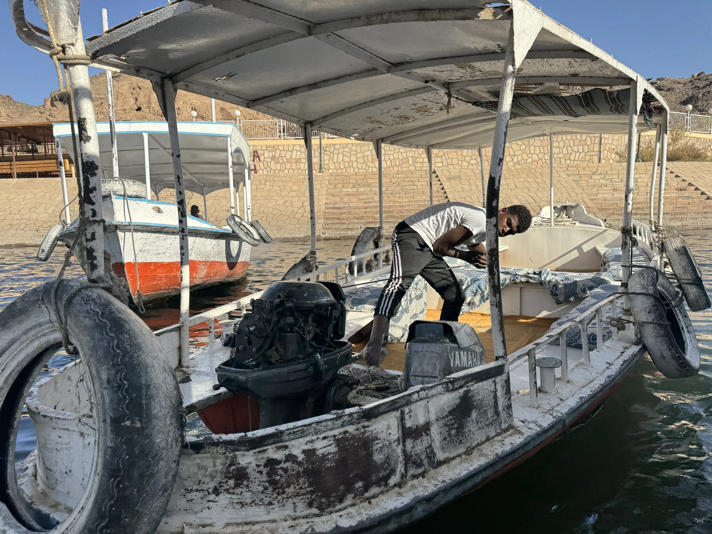 A man on a boat adjusting equipment, with white and blue painted boats around, in a desert landscape with rocky hills in the background.