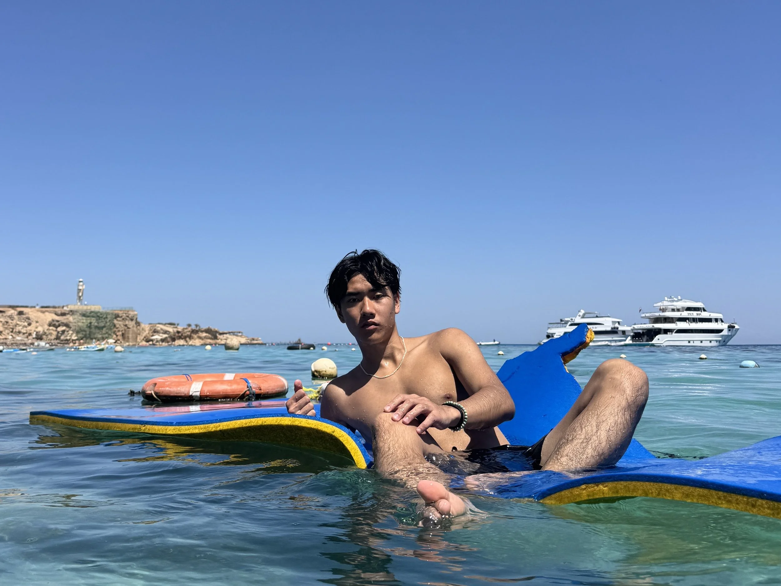Young man relaxing on a paddleboard in clear blue water with boats and a rocky coastline in the background under a bright blue sky.