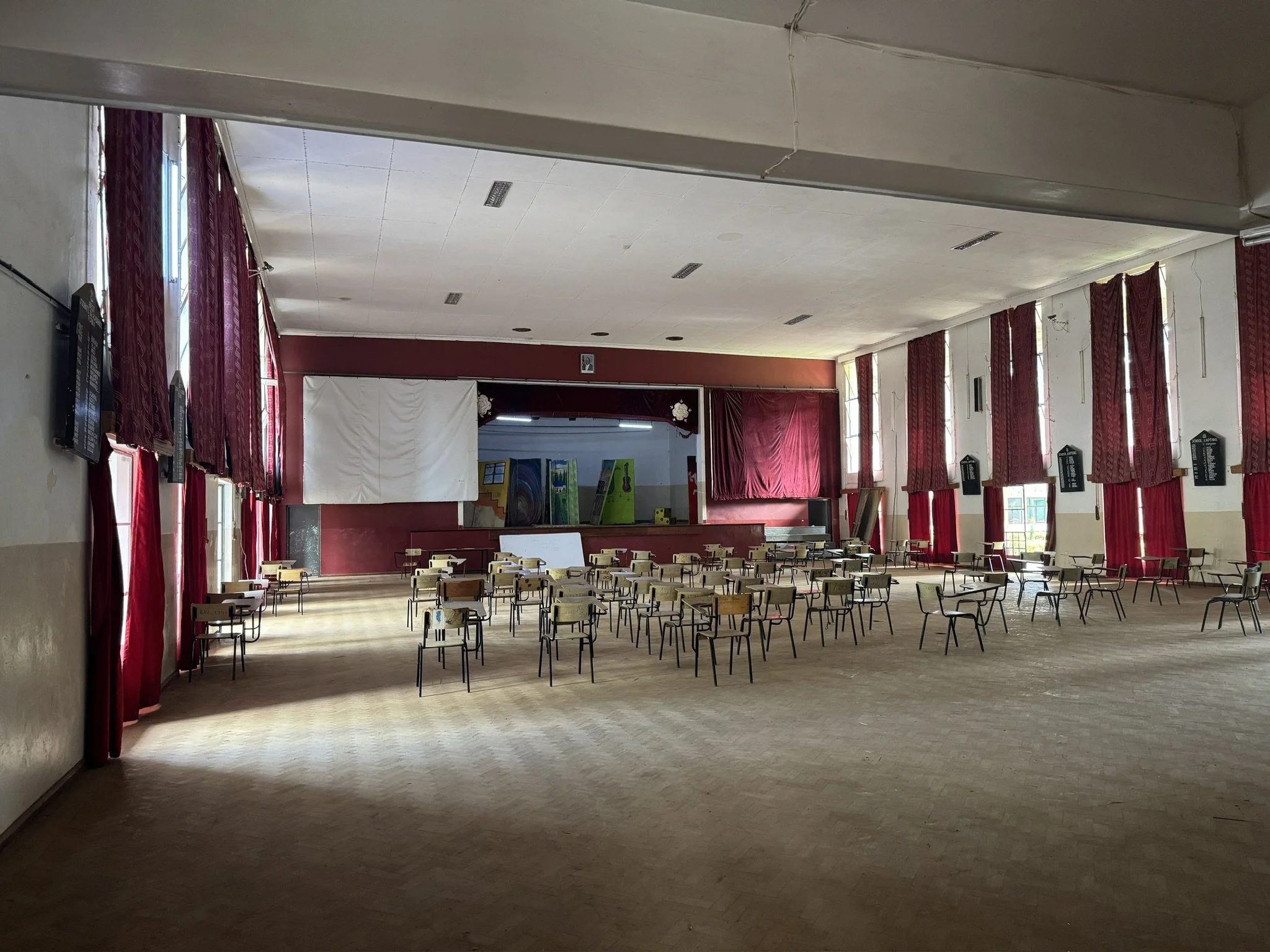 An empty auditorium or hall with rows of chairs, red curtains along the windows, and some artwork and paint on display at the back of the room.