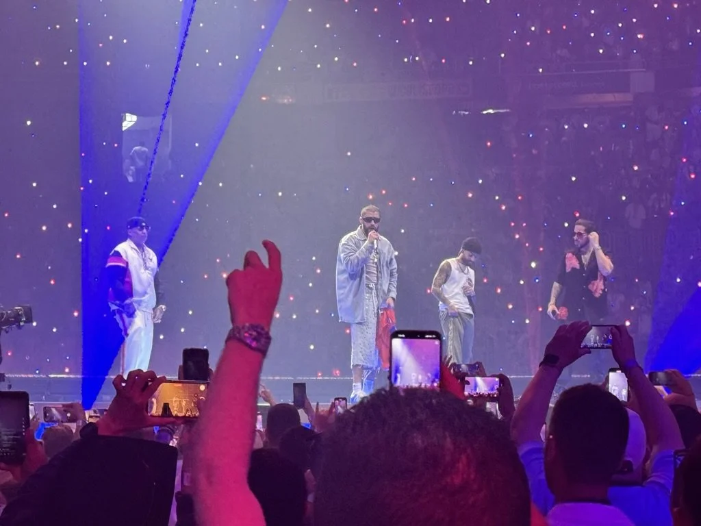 Four performers on stage during a concert with a colorful light display behind them. Audience members are taking photos and videos with their phones.