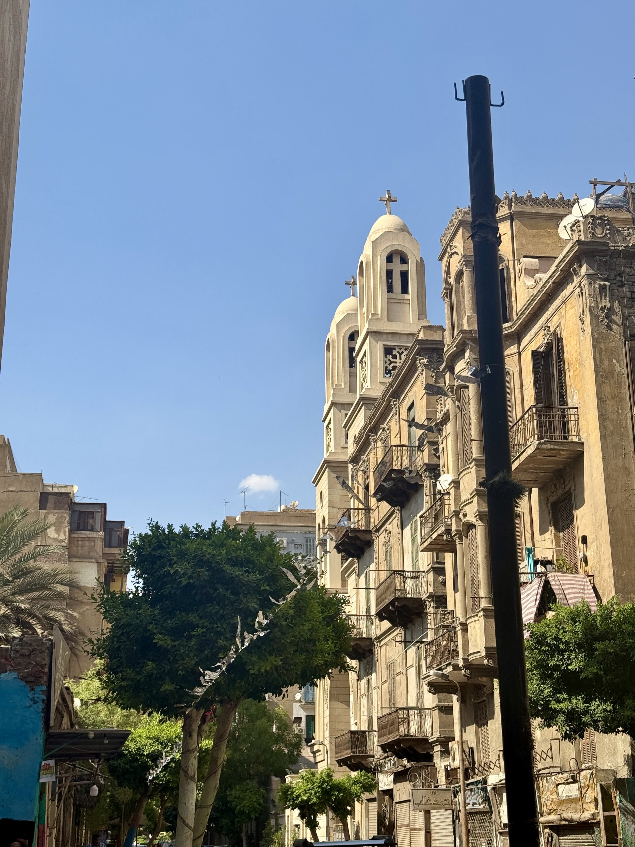 Street view showing a tall beige building with ornate balconies and a church with bell towers topped with crosses. Trees and a blue sky fill the scene.