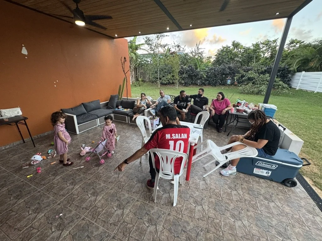 A group of people sitting under a covered patio, with some children playing on the tiled floor, and a grassy yard with trees in the background.
