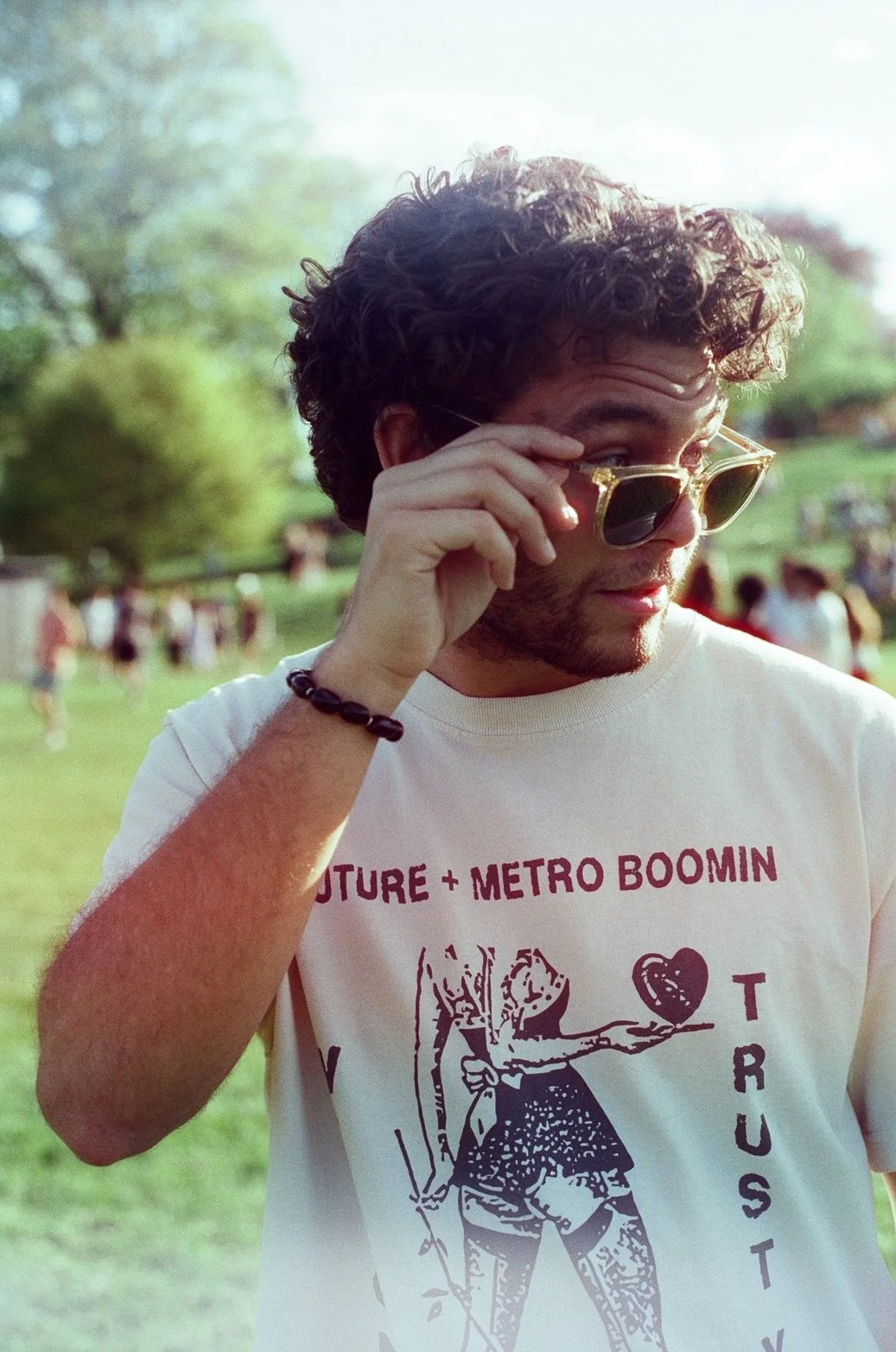 A young man with curly hair and a beard wearing sunglasses and a white T-shirt with graphics and text. He is outdoors in a park, holding his sunglasses with one hand and looking slightly downward.