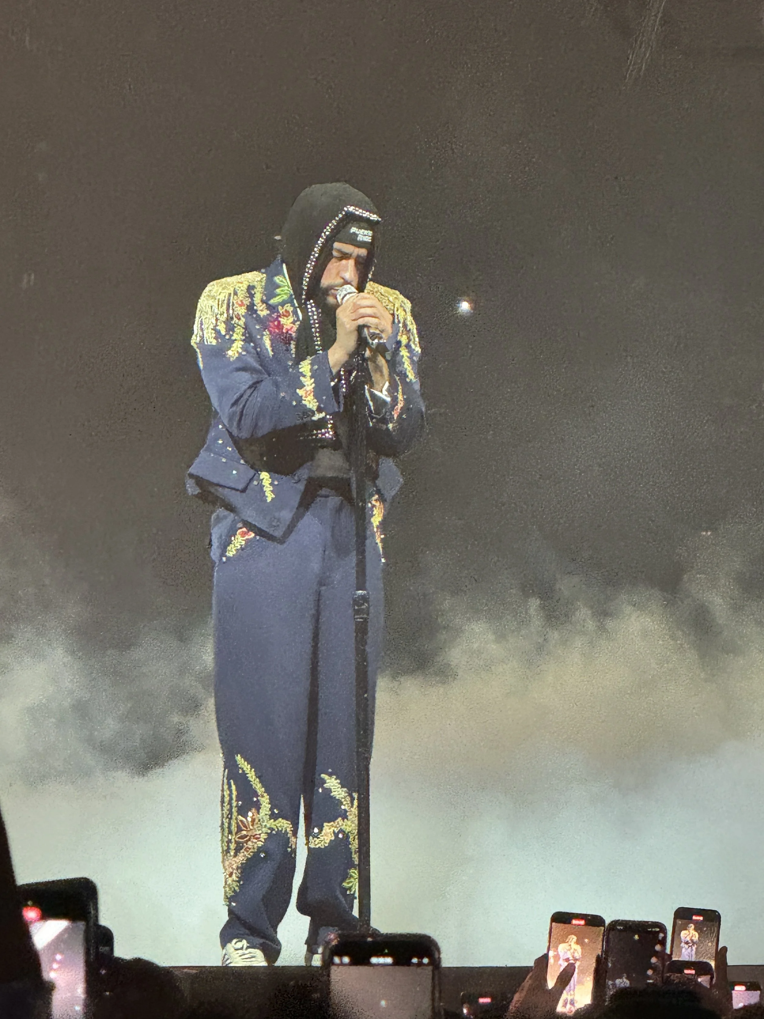 A performer on stage wearing a black hooded jacket with gold and floral embroidery, and a black beanie, singing into a microphone. Audience members are taking photos or videos with their smartphones.