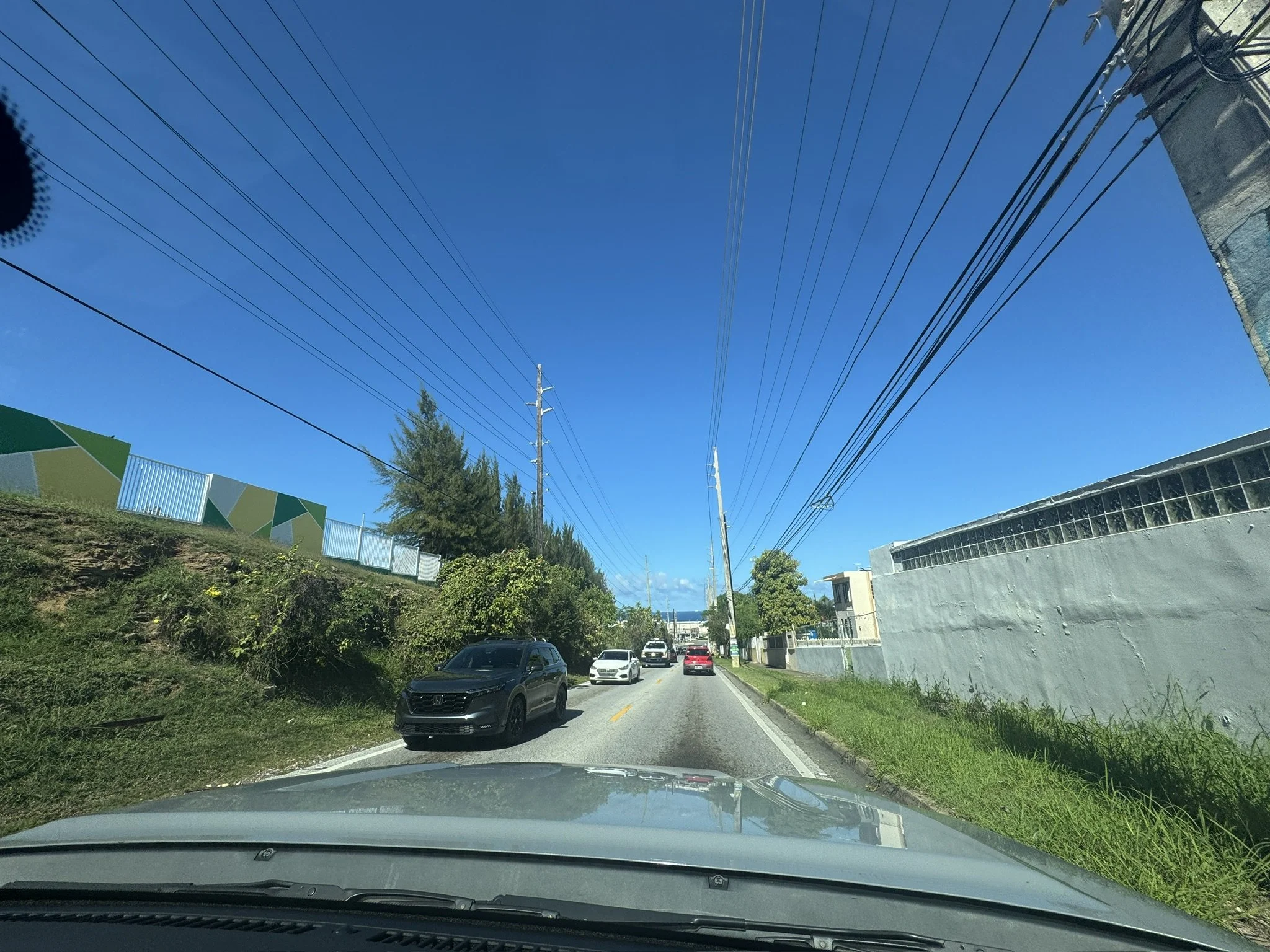 A view from a car during a road trip showing cars driving on a street with a fence and trees on the sides, power lines overhead, and a clear blue sky.