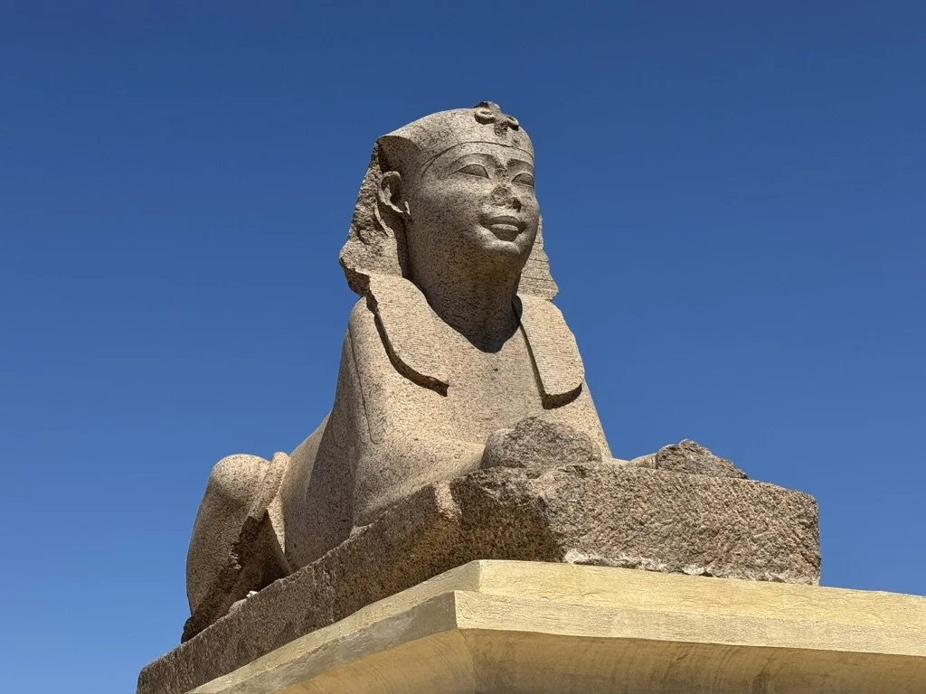 Stone statue of a smiling woman with Egyptian headdress, set against a blue sky.