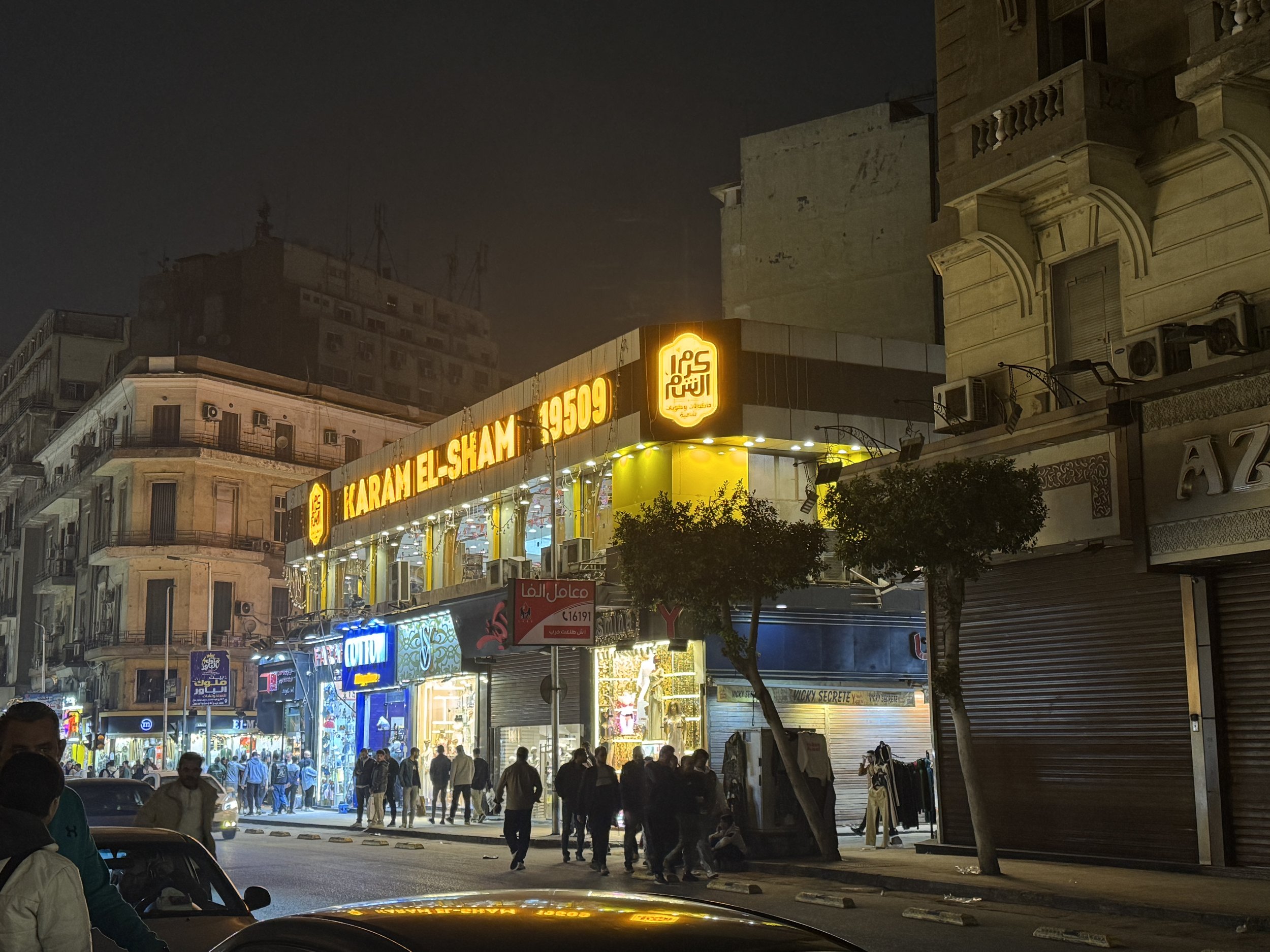 Night scene of a busy city street with illuminated storefronts and signs, and a crowd of people walking along the sidewalk.