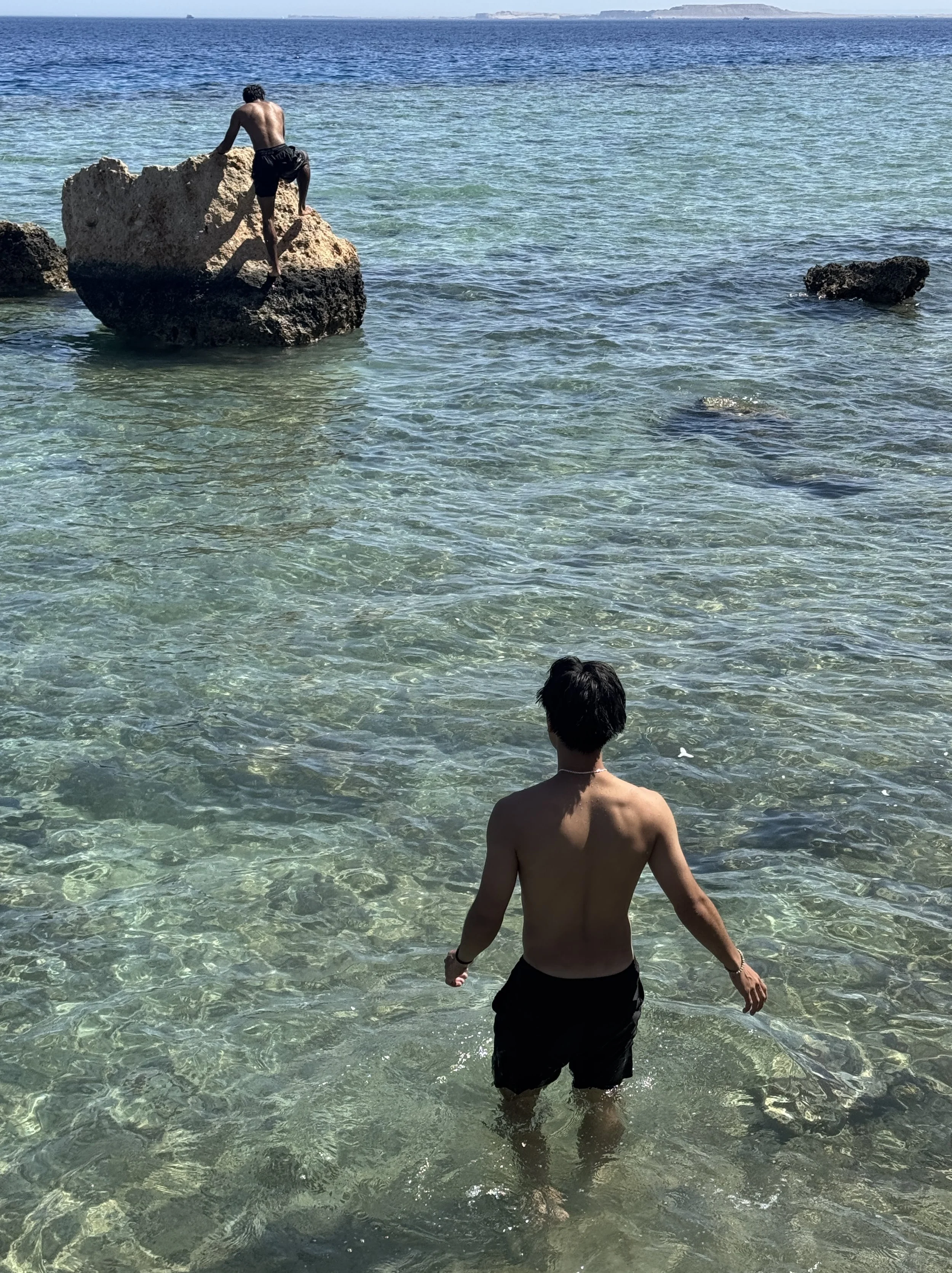 A boy standing in shallow clear water at the beach, wearing black shorts, facing away, with two large rocks and a person climbing on one of them in the background.