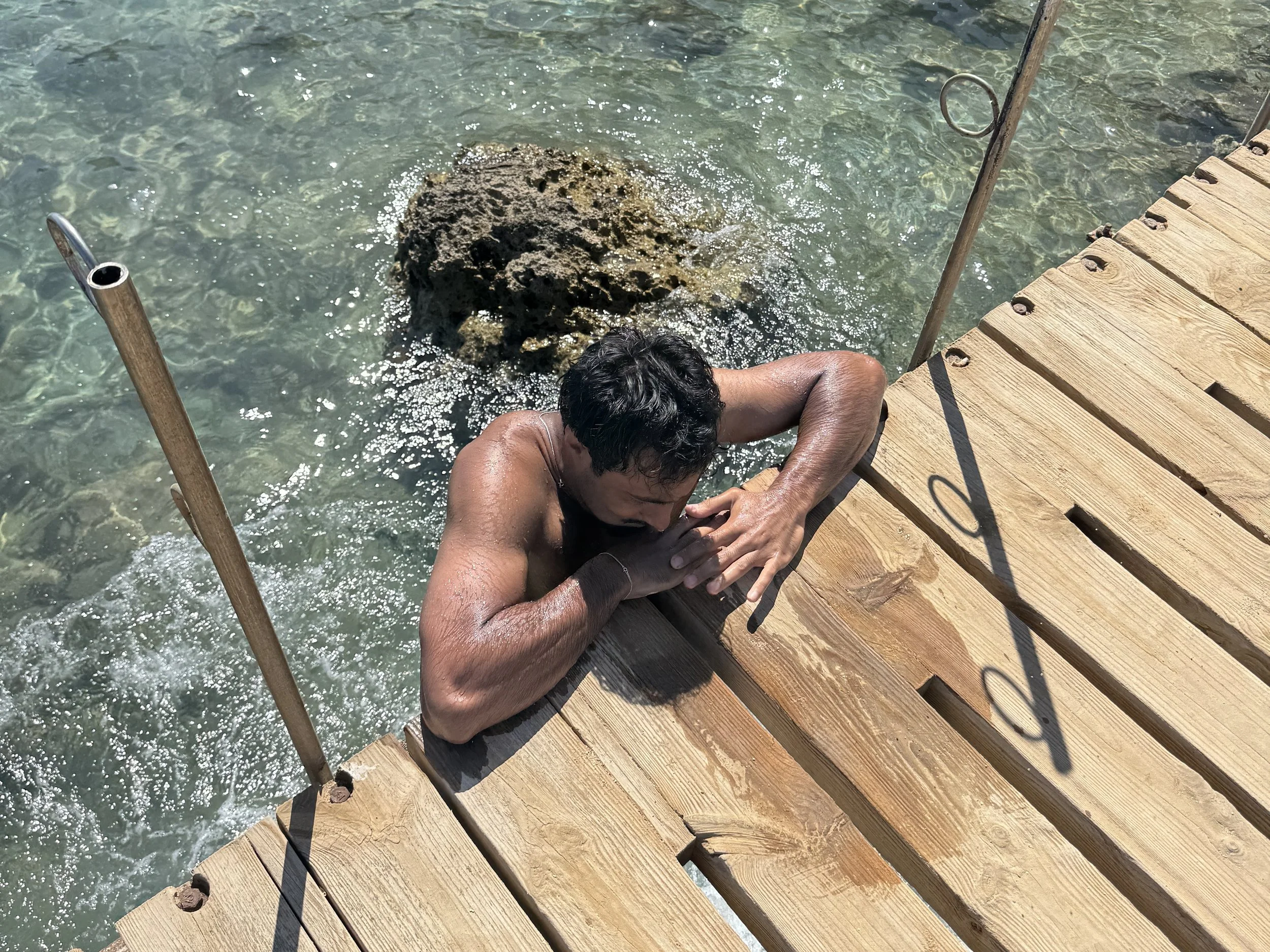 A person with dark hair is climbing up a wooden dock into the water, holding onto the dock's edge. The water is clear and the dock has metal handrails.