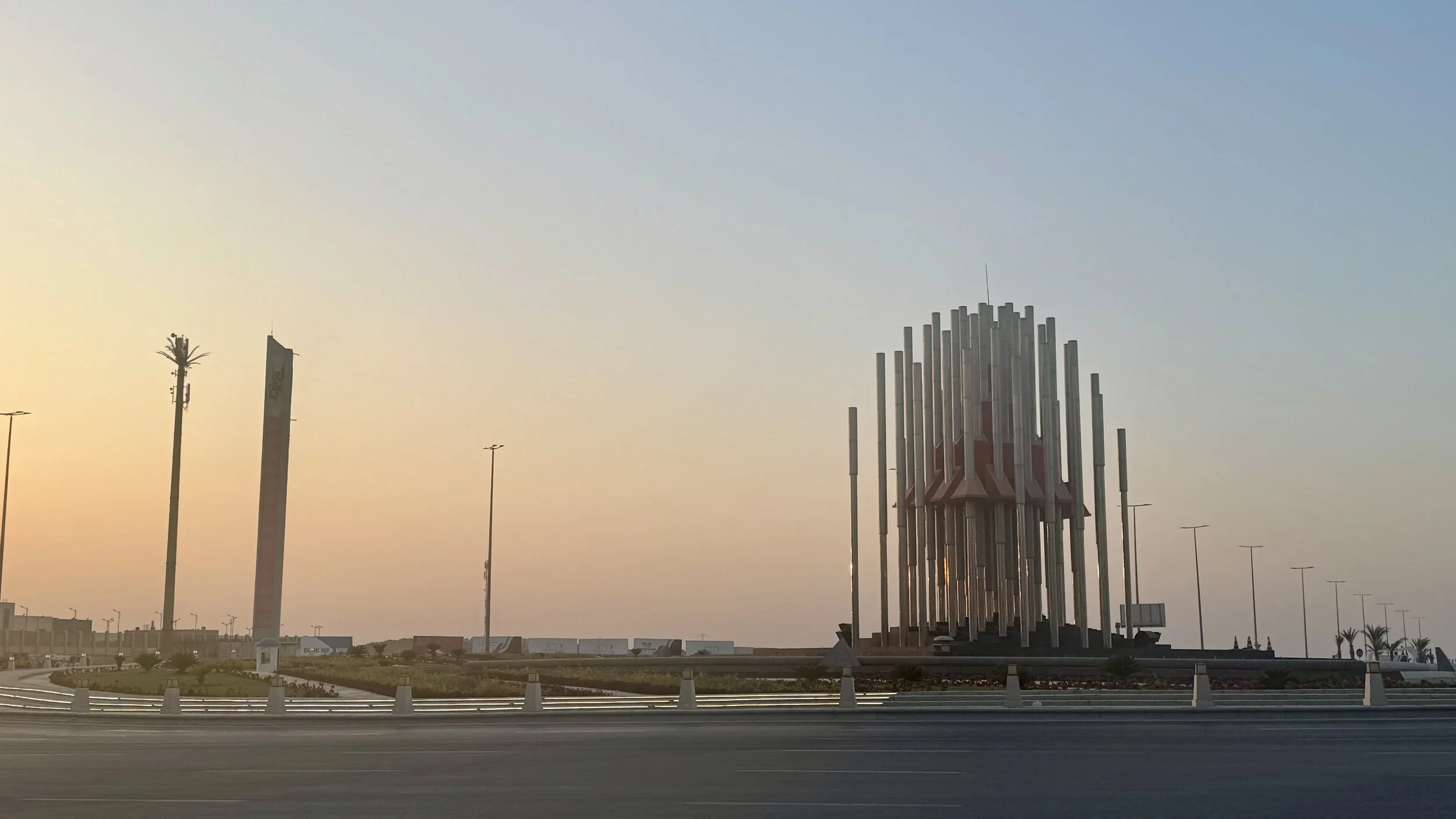 A monument with tall vertical metallic pillars, a large flagpole with a flag to the left, and a clear sky at sunset.