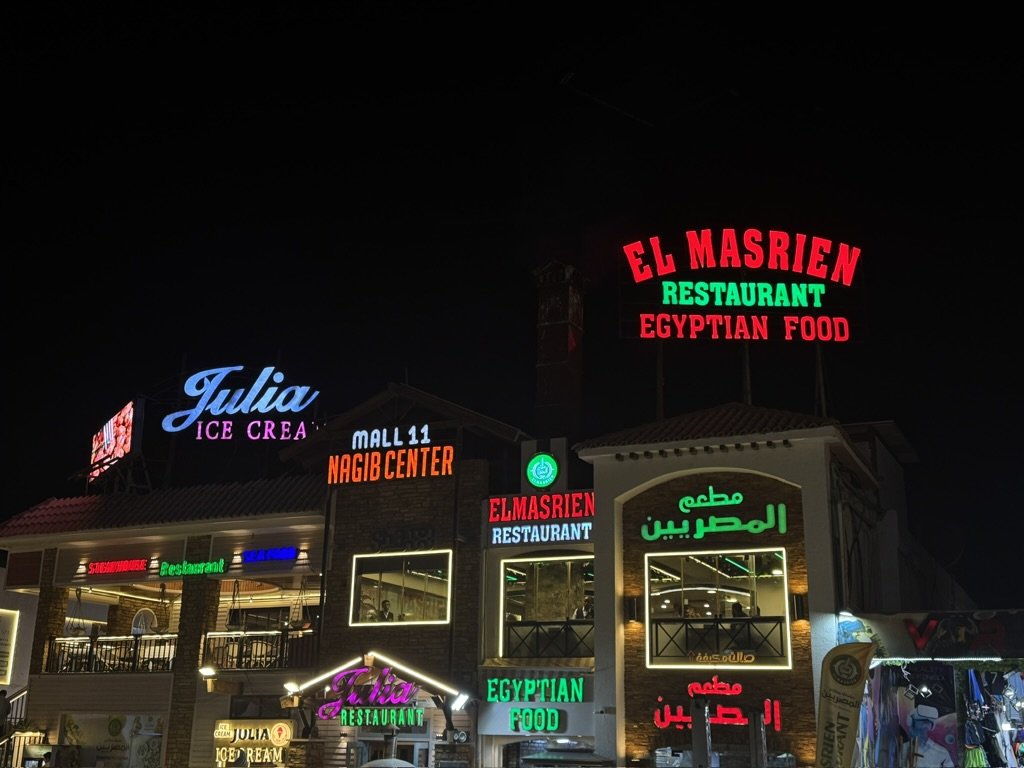Night view of a building with colorful neon signs advertising restaurants, including 'El Masrien,' 'Julia Ice Cream,' and 'El Masrien Egyptian Food,' with Arabic script also visible.